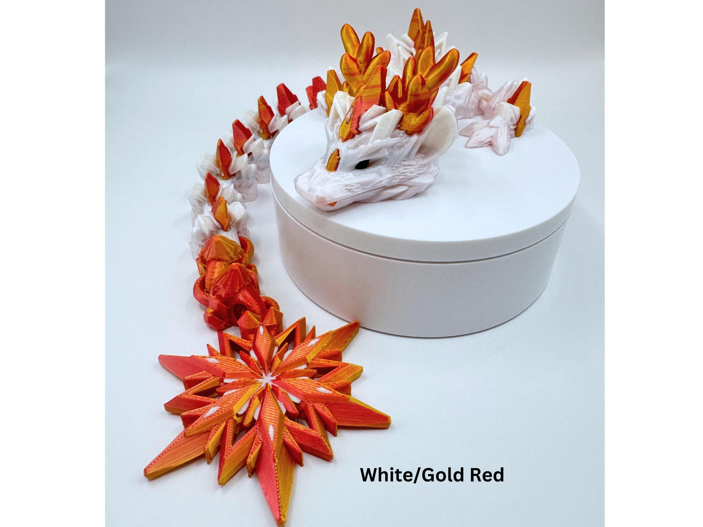 A white and gold red winter dragon figurine with a red snowflake tail. The dragon is laying on a white round box with a red snowflake at the end of the tail. The text 'White/Gold Red' is below the star.
