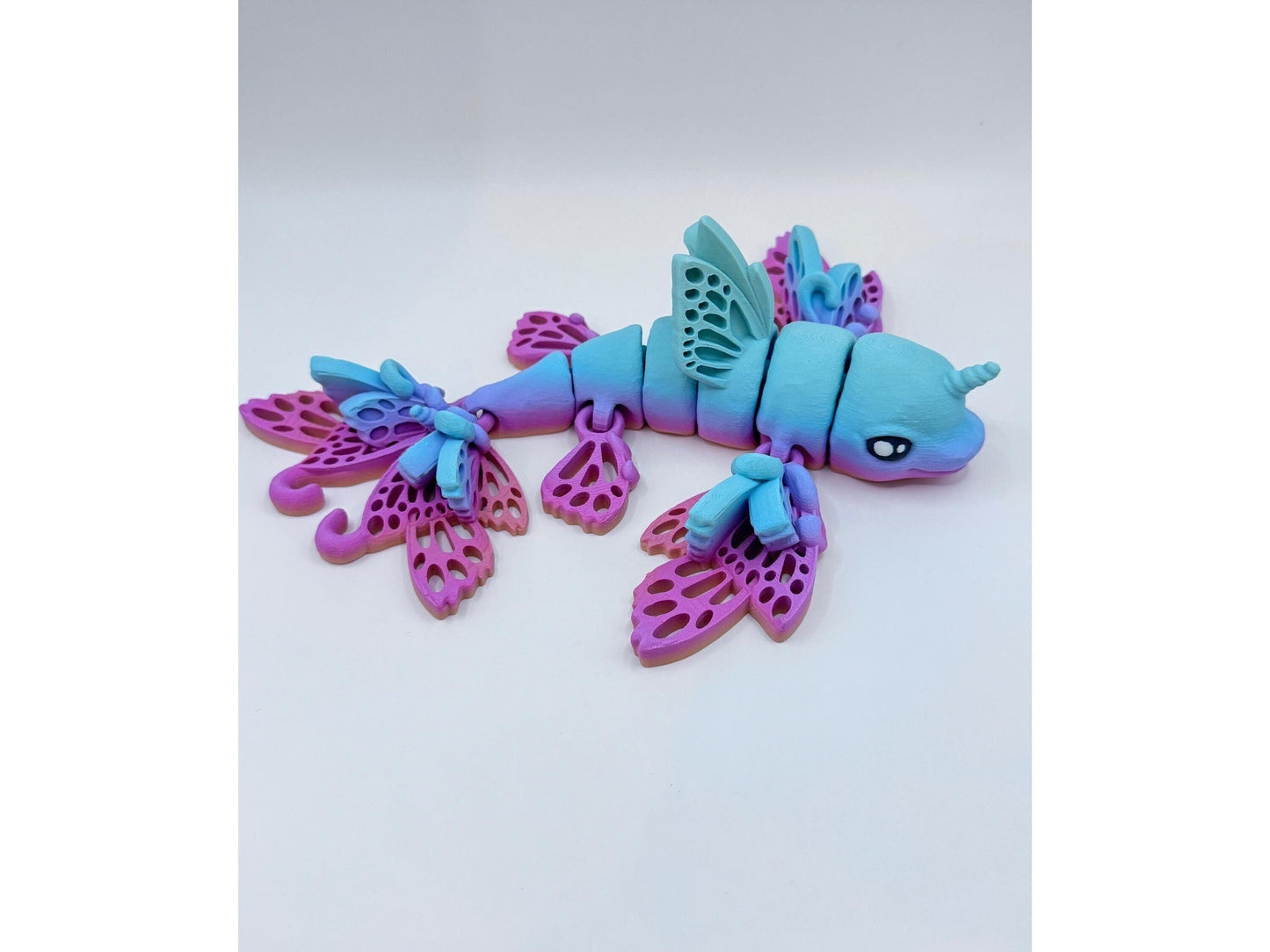 A 3D-printed, articulated dolphin fidget with butterfly wings in a mesmerizing blue and pink rainbow color features a unicorn horn adorning its head on a white background.