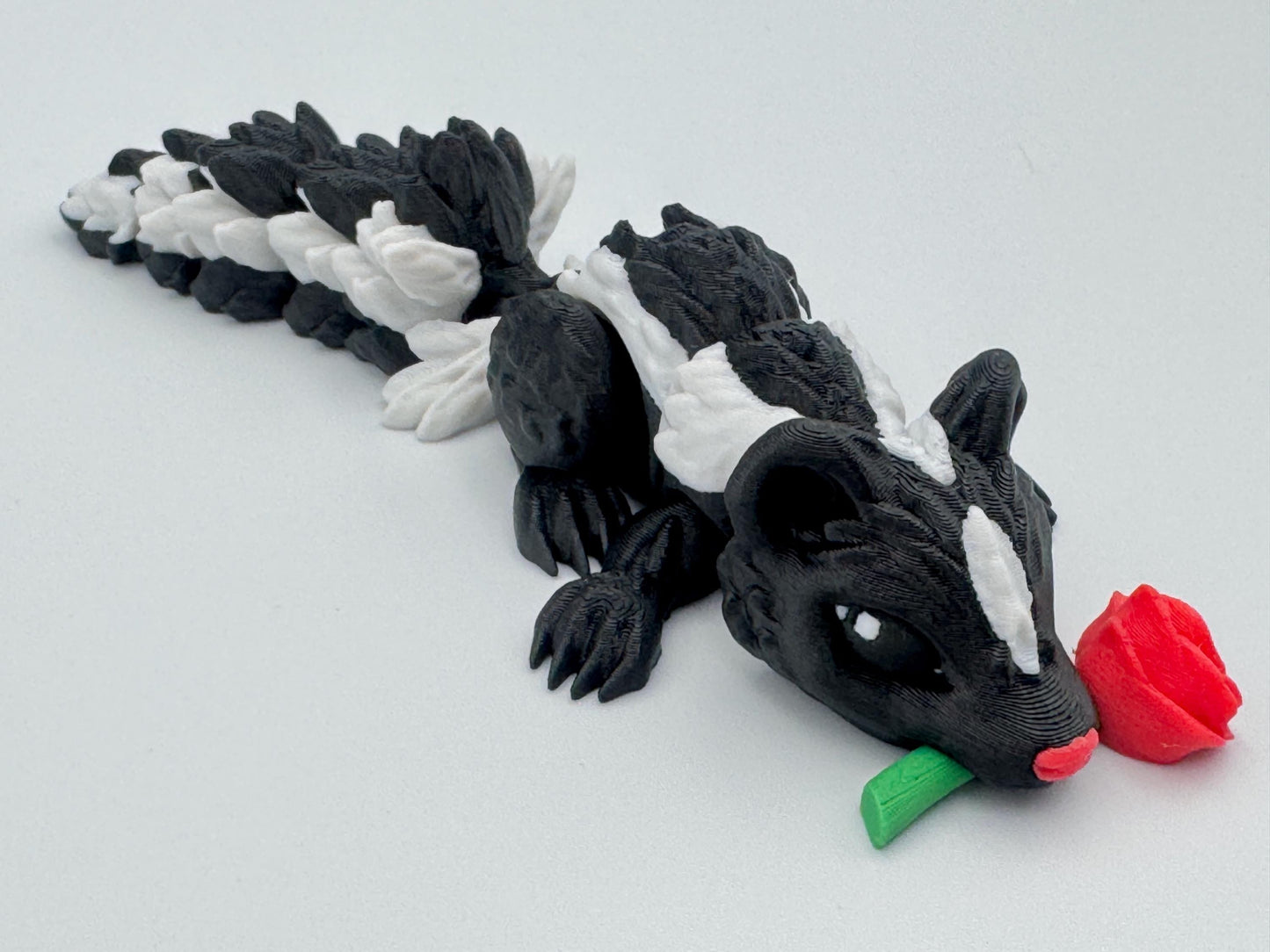 3D printed articulated Rose Skunk figurine featuring a black and white skunk holding a red rose in its mouth. The skunk has a flexible, segmented body and detailed texture, designed as a playful and charming Valentine’s Day decoration or collectible.