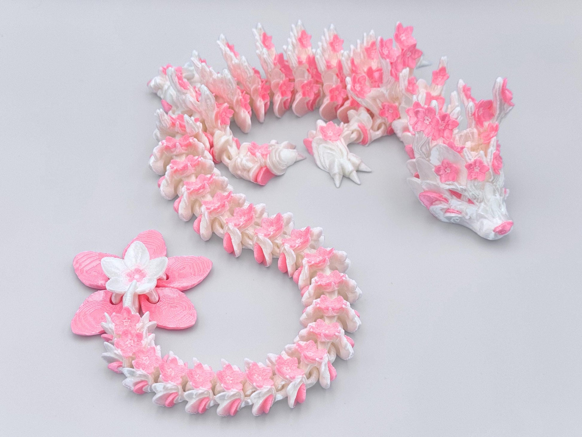 White and pink silk Cherry Blossom dragon figurine with articulated body laid in an S-curve. The 3D printed design highlights floral accents, detailed blossoms, and a large pink and white flower tail.