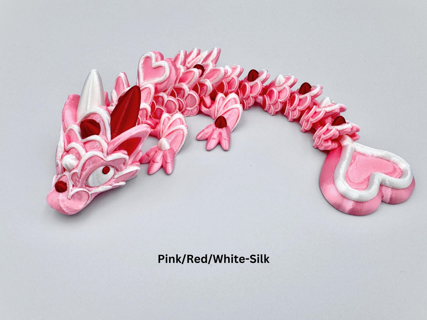 3D-printed articulated Baby Heart Cookie Dragon with a pink silk body, white frosting-like patterns, and vibrant red accents. Features intricate details, a fully flexible design, and is perfect as a unique Valentines Day gift or playful decoration.