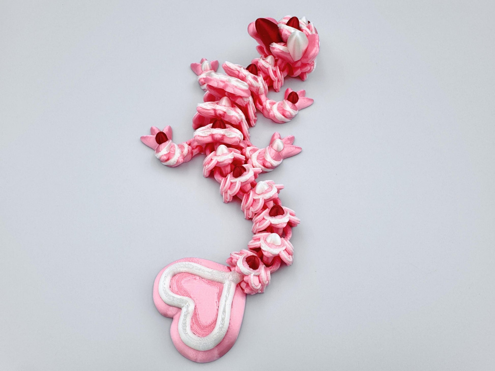 3D-printed articulated Baby Heart Cookie Dragon with a pink silk body, white frosting-like patterns, and vibrant red accents. Features intricate details, a fully flexible design, and is perfect as a unique Valentines Day gift or playful decoration.