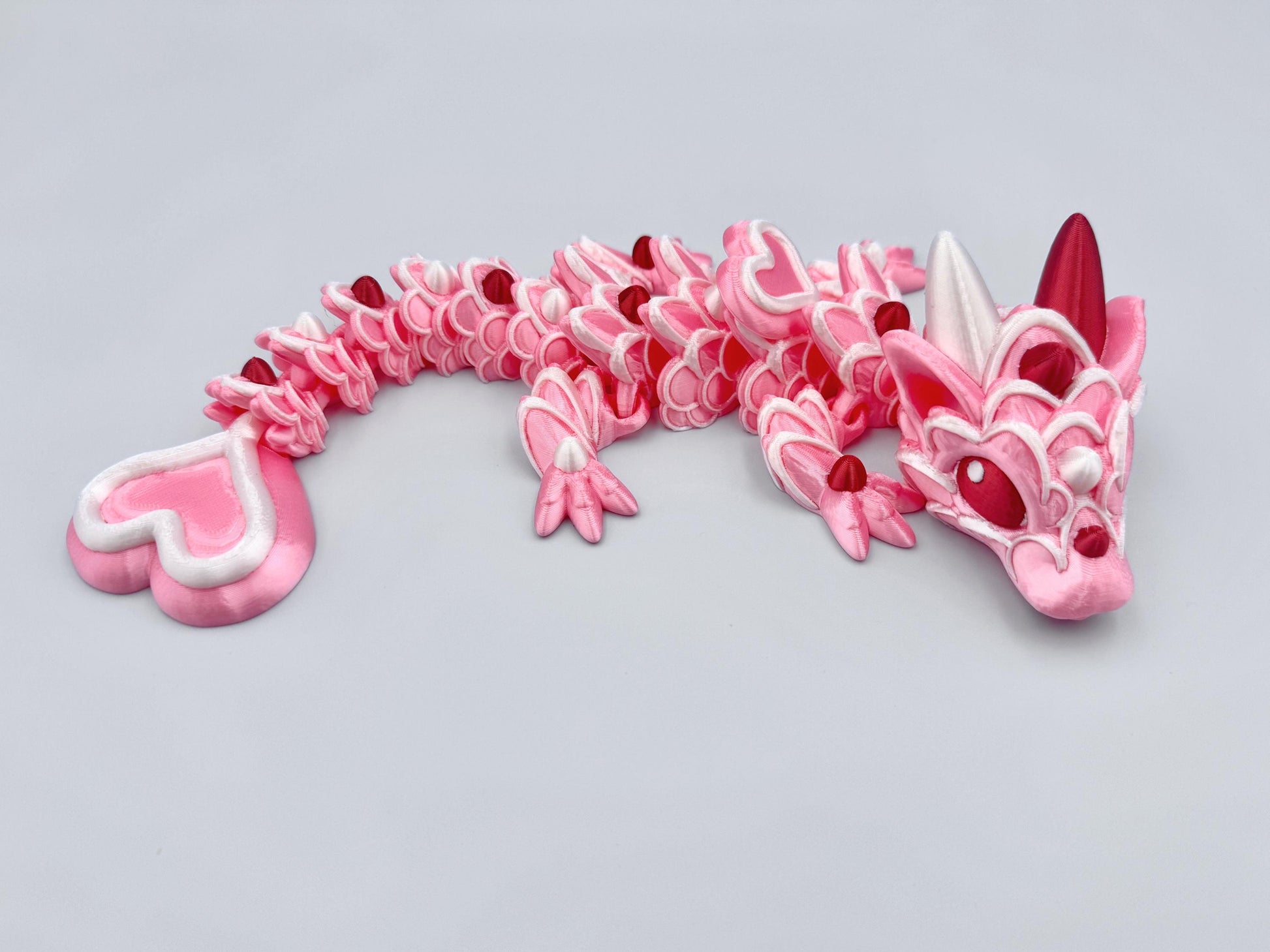 3D-printed articulated Baby Heart Cookie Dragon with a pink silk body, white frosting-like patterns, and vibrant red accents. Features intricate details, a fully flexible design, and is perfect as a unique Valentines Day gift or playful decoration.