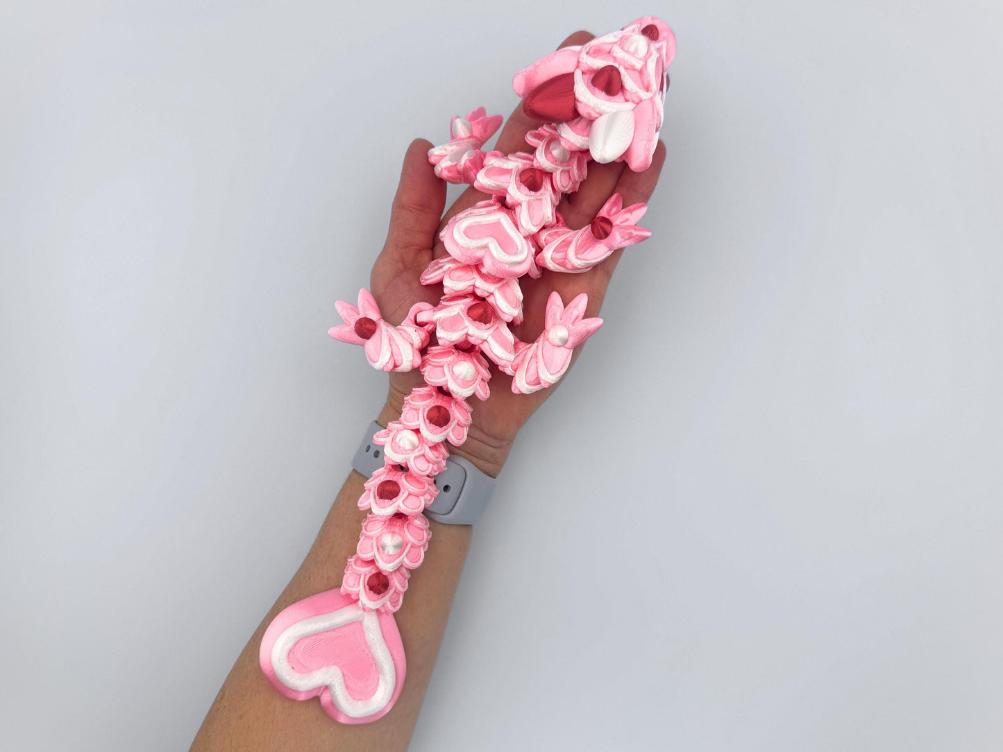 3D-printed articulated Baby Heart Cookie Dragon with a pink silk body, white frosting like patterns, and vibrant red accents. Features intricate details, a fully flexible design, and is perfect as a unique Valentines Day gift or playful decoration.