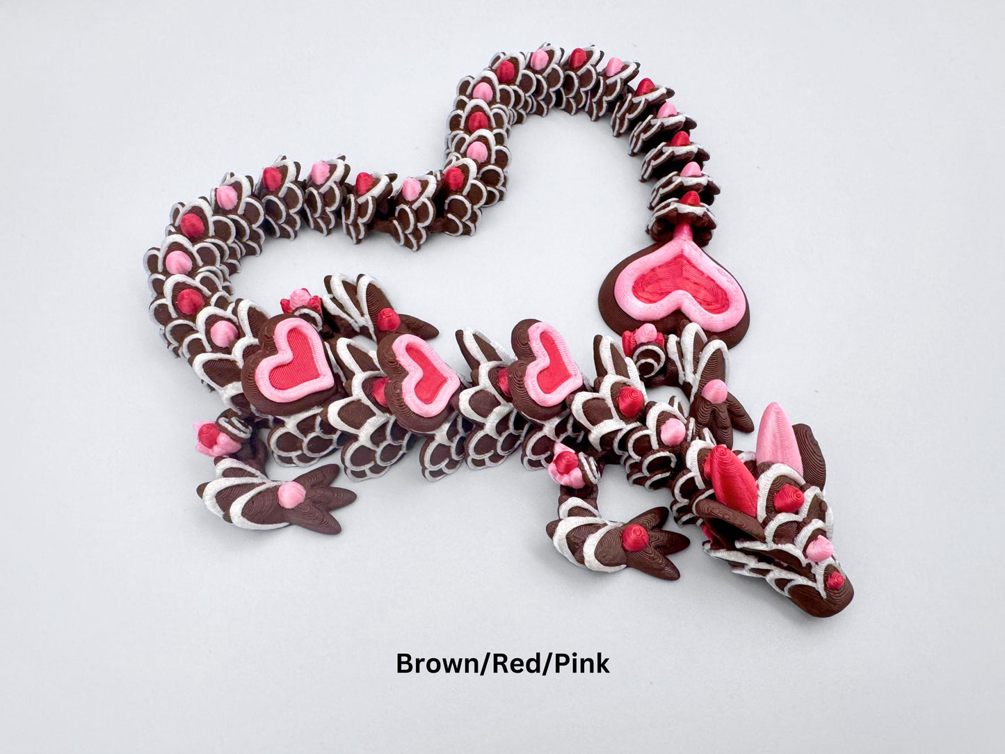 3D-printed articulated heart dragon cookie inspired fidget toy, with dark chocolate brown body, silk pink and silk red candy accents, white icing-like patterns and red hears with pink outline. Features intricate details and a fully flexible design.