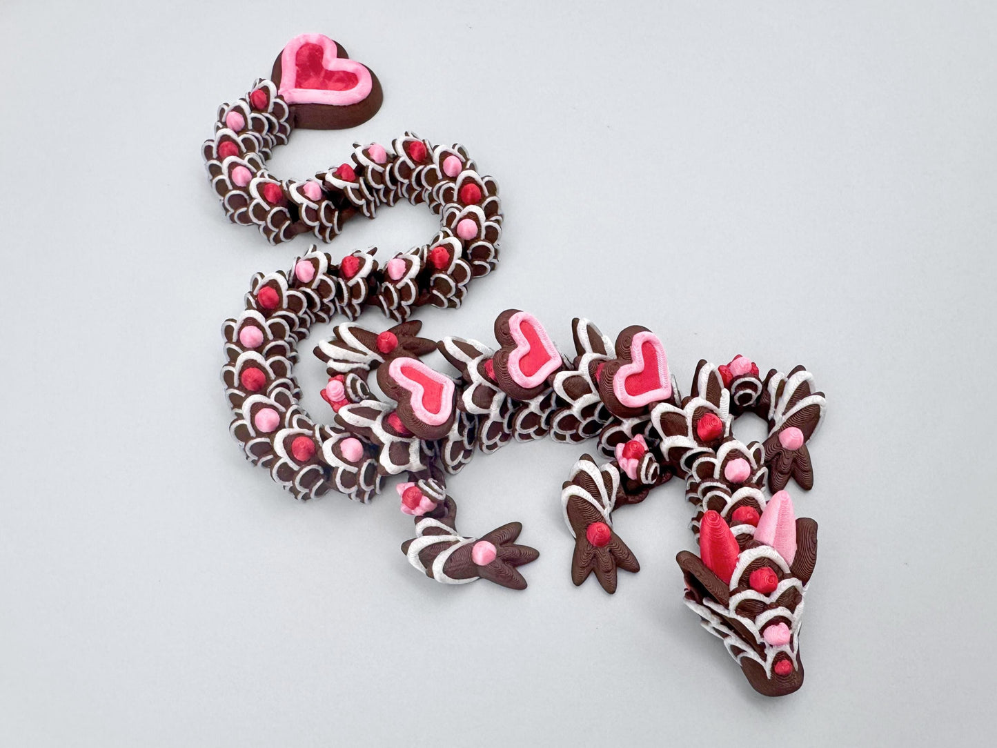 3D-printed articulated heart dragon cookie inspired fidget toy, with dark chocolate brown body, silk pink and silk red candy accents, white icing-like patterns and red hears with pink outline. Features intricate details and a fully flexible design.