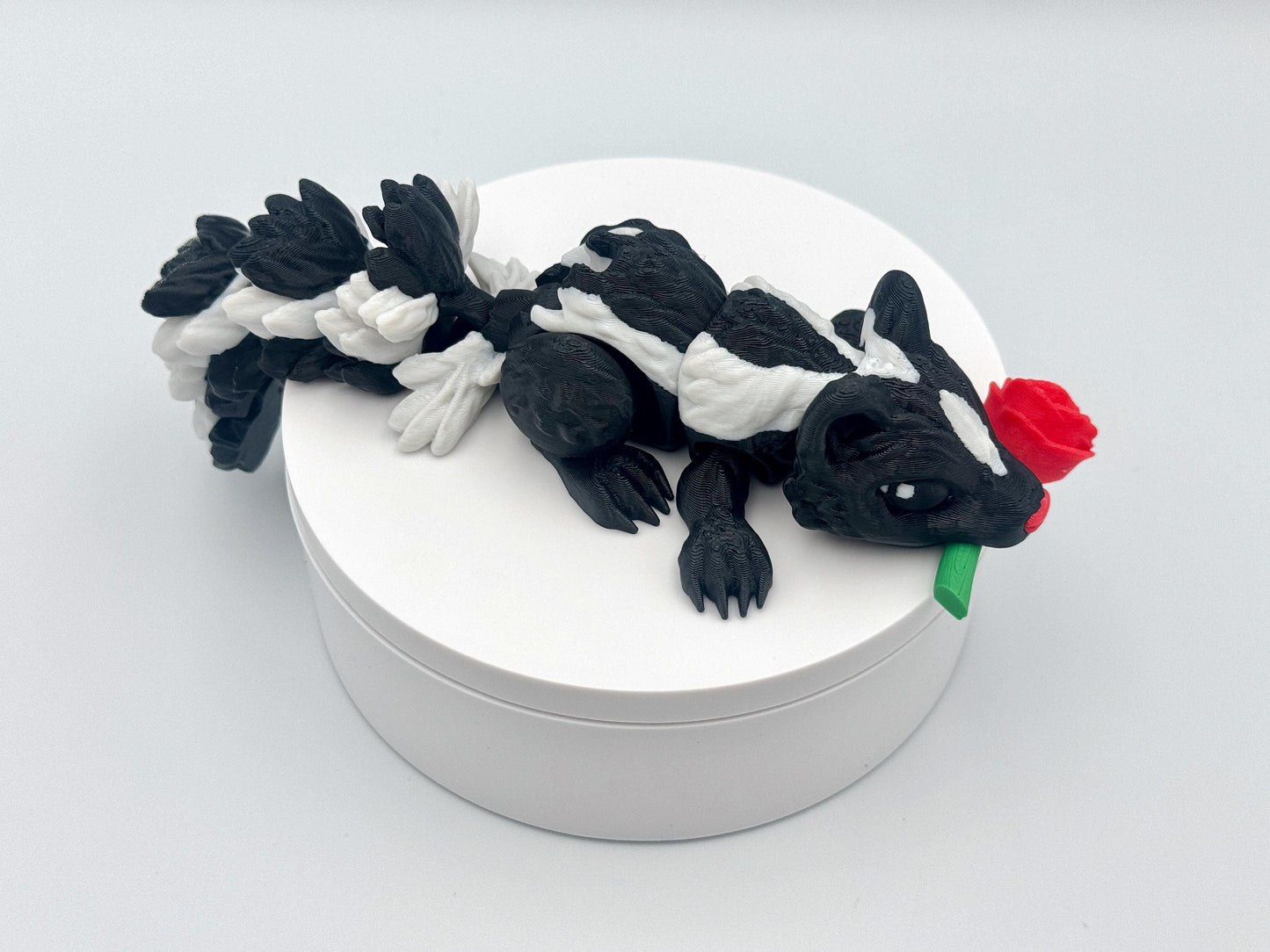 3D printed articulated Rose Skunk figurine featuring a black and white skunk holding a red rose in its mouth. The skunk has a flexible, segmented body and detailed texture, designed as a playful and charming Valentine’s Day decoration or collectible.