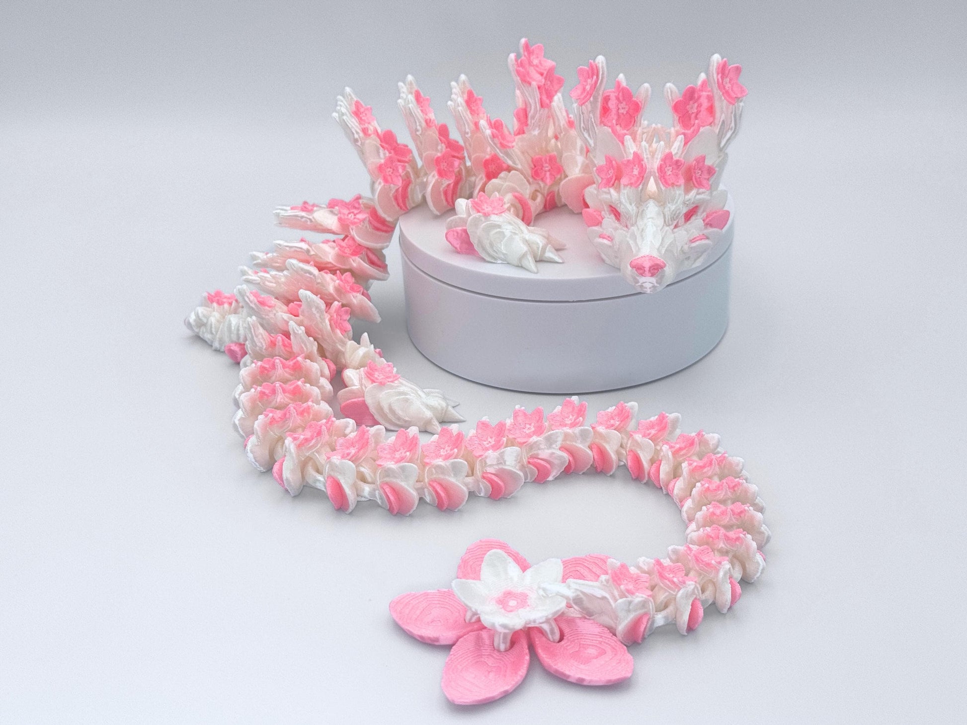 3D printed Cherry Blossom articulated dragon in white and pink silk, posed on a round display stand. The figurine has layered flower details along the body, flexible joints and a large blossom tail tip.