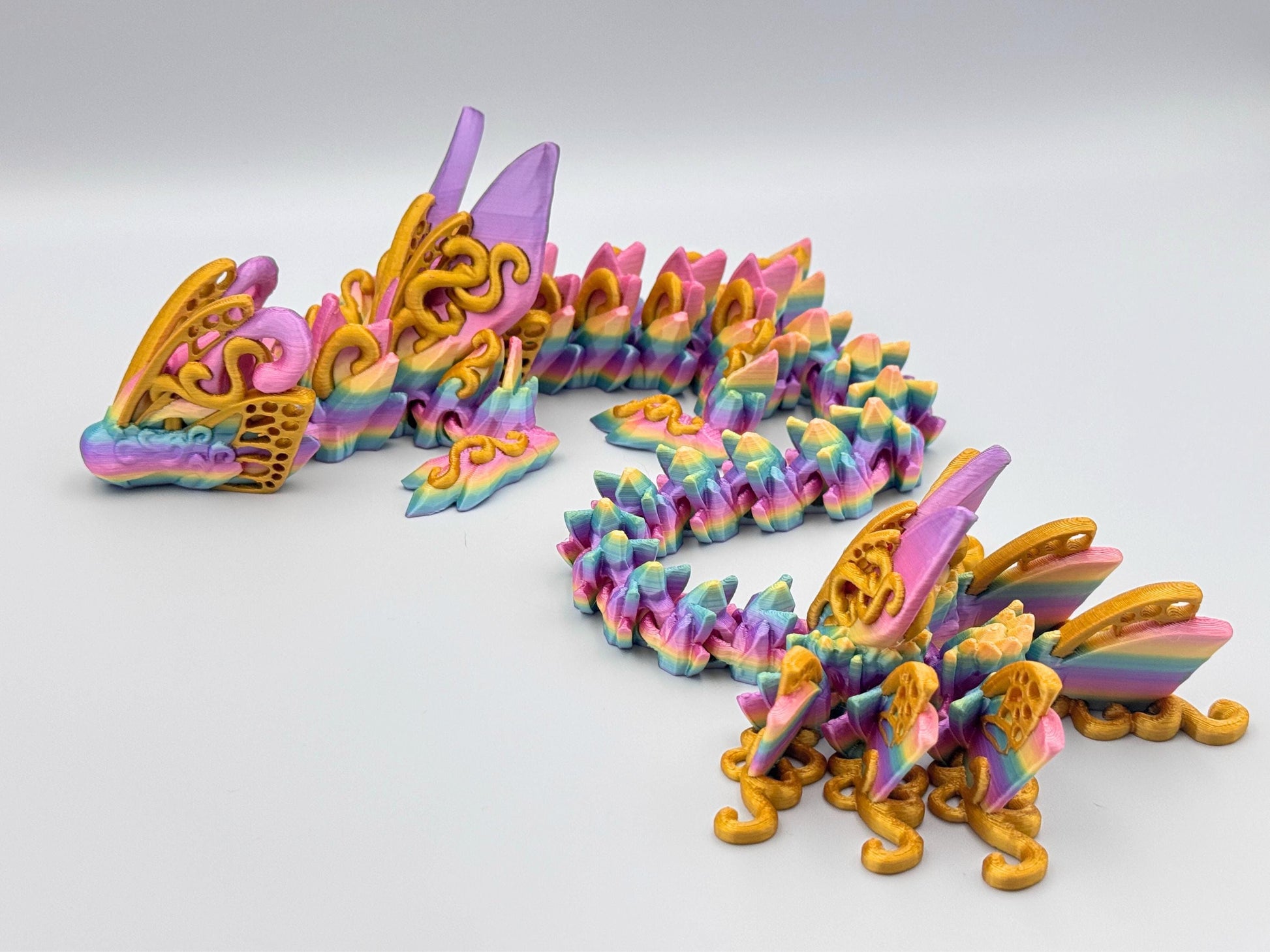3D printed butterfly dragon figurine in rainbow filament with gold accents along the body, curled on a white surface with articulated segments and sculpted texture.