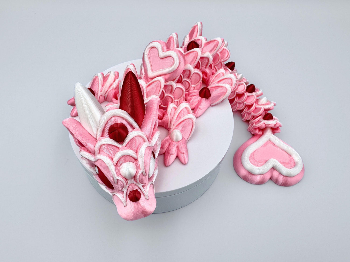 3D-printed articulated Baby Heart Cookie Dragon with a pink silk body, white frosting-like patterns, and vibrant red accents. Features intricate details, a fully flexible design, and is perfect as a unique Valentines Day gift or playful decoration.