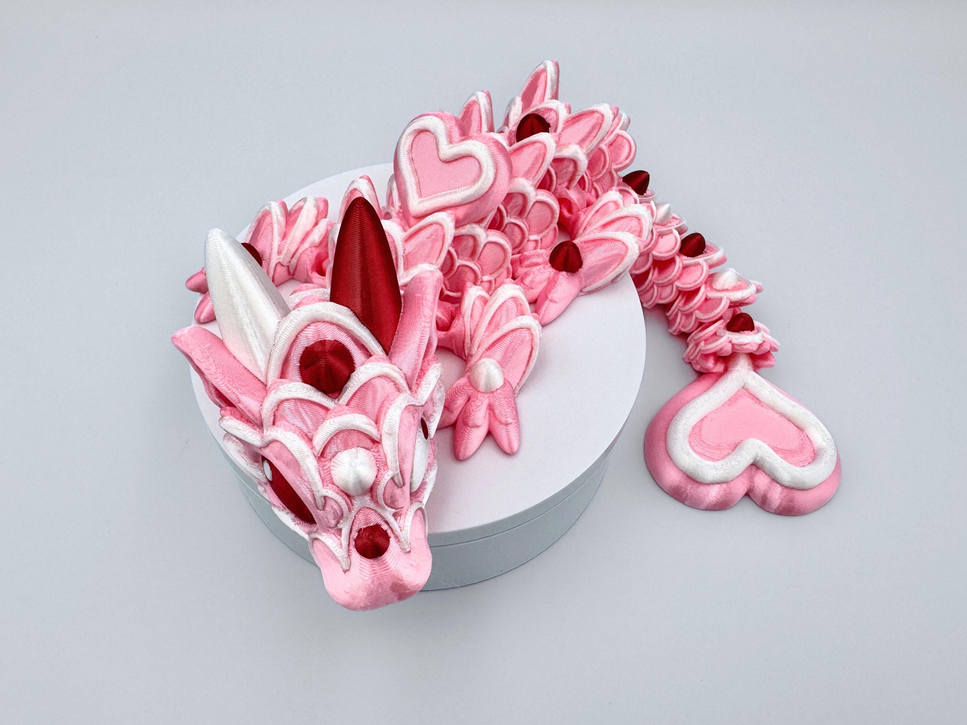 3D-printed articulated Baby Heart Cookie Dragon with a pink silk body, white frosting-like patterns, and vibrant red accents. Features intricate details, a fully flexible design, and is perfect as a unique Valentines Day gift or playful decoration.