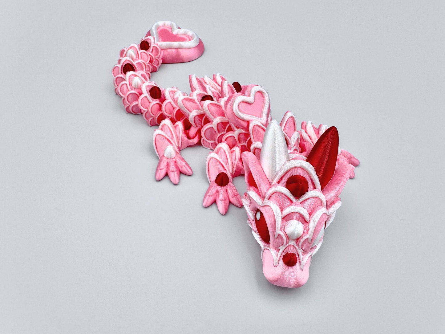 3D-printed articulated Baby Heart Cookie Dragon with a pink silk body, white frosting-like patterns, and vibrant red accents. Features intricate details, a fully flexible design, and is perfect as a unique Valentines Day gift or playful decoration.