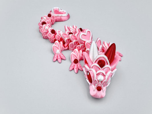 3D-printed articulated Baby Heart Cookie Dragon with a pink silk body, white frosting-like patterns, and vibrant red accents. Features intricate details, a fully flexible design, and is perfect as a unique Valentines Day gift or playful decoration.