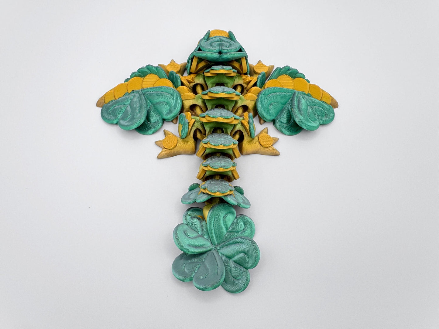 A 3D printed baby clover dragon figurine displayed flat on a white background, viewed directly from above. The dragon’s limbs are stretched outward, and the full body is covered with clover leaf shapes, including the wings, spine, tail, head, and feet. The symmetrical layout shows how the clover leaves form the dragon’s structure in repeating patterns. The head is at the bottom center and the tail tapers toward the top of the image.