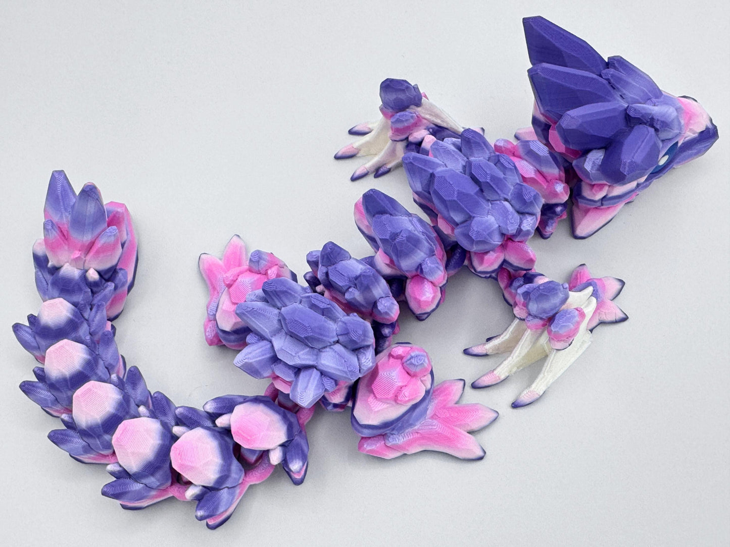 3D-printed Gemstone Baby Dragon fidget toy with an articulated body, featuring vibrant purple, pink, and white crystal-like scales. The segmented design allows flexible movement, while the intricate faceted details mimic the look of precious gemstones, creating a striking and mystical aesthetic.