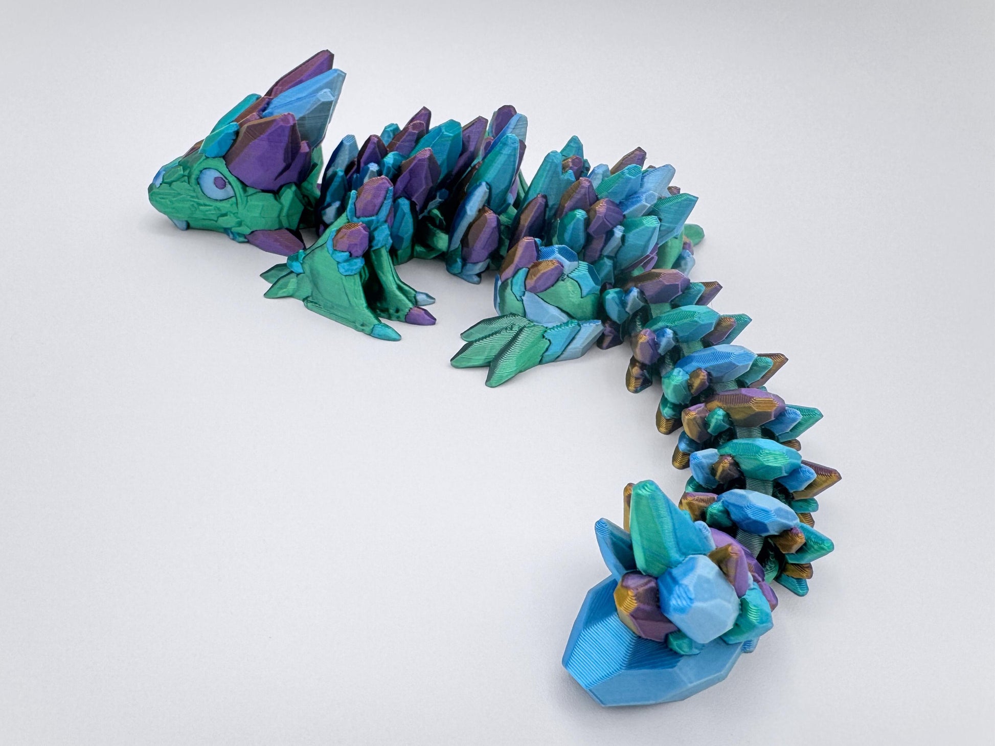 3D-printed Gemstone Baby Dragon fidget toy with an articulated body, featuring green, purple, gold and blue in silver and gold crystal scales. The segmented design allows flexible movement, while the intricate faceted details mimic the look of precious gemstones, creating a striking and mystical aesthetic.