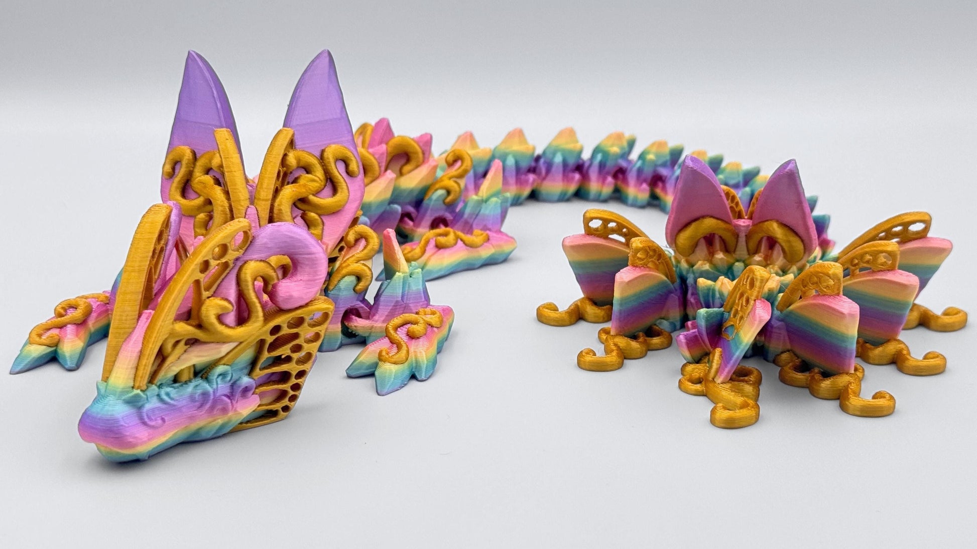 3D printed butterfly dragon figurine in rainbow filament with gold accents and details, shown curled on a white surface with articulated segments and sculpted texture.