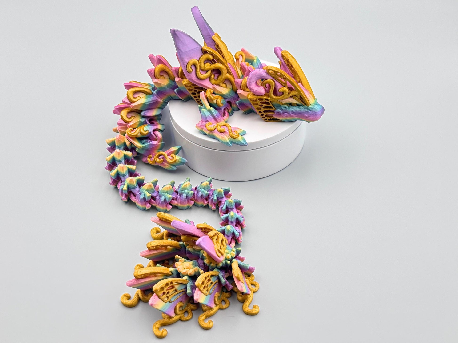 3D printed butterfly dragon figurine in rainbow filament with gold accents and details, shown curled on a white stand with articulated segments and sculpted texture.
