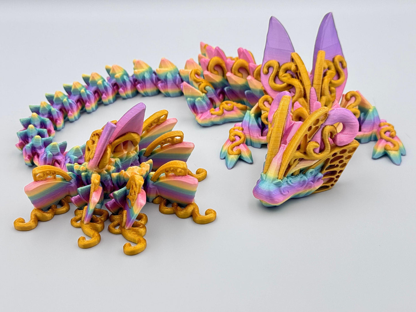 3D printed butterfly dragon figurine in rainbow filament with gold accents along the body, curled on a white surface with articulated segments and sculpted texture.