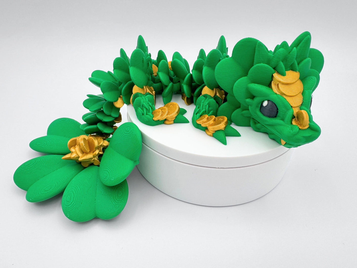This 3D Printed Clover Dragon green and gold articulated figurine with a clover 4 leaf design along its body.  Used as desk decor for seasonal gift for St. Patrick’s Day. The dragon is lying on a white surface.