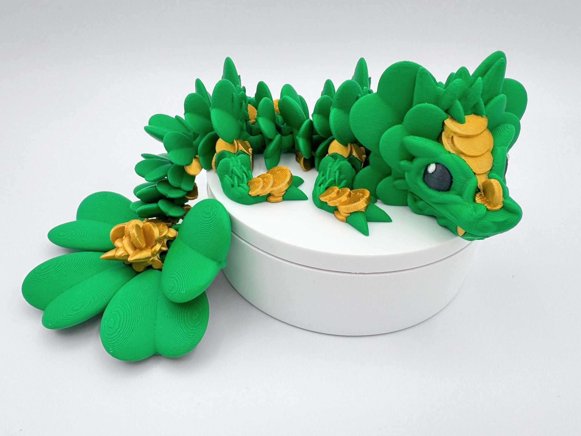 This 3D Printed Clover Dragon green and gold articulated figurine with a clover 4 leaf design along its body.  Used as desk decor for seasonal gift for St. Patrick’s Day. The dragon is lying on a white surface.