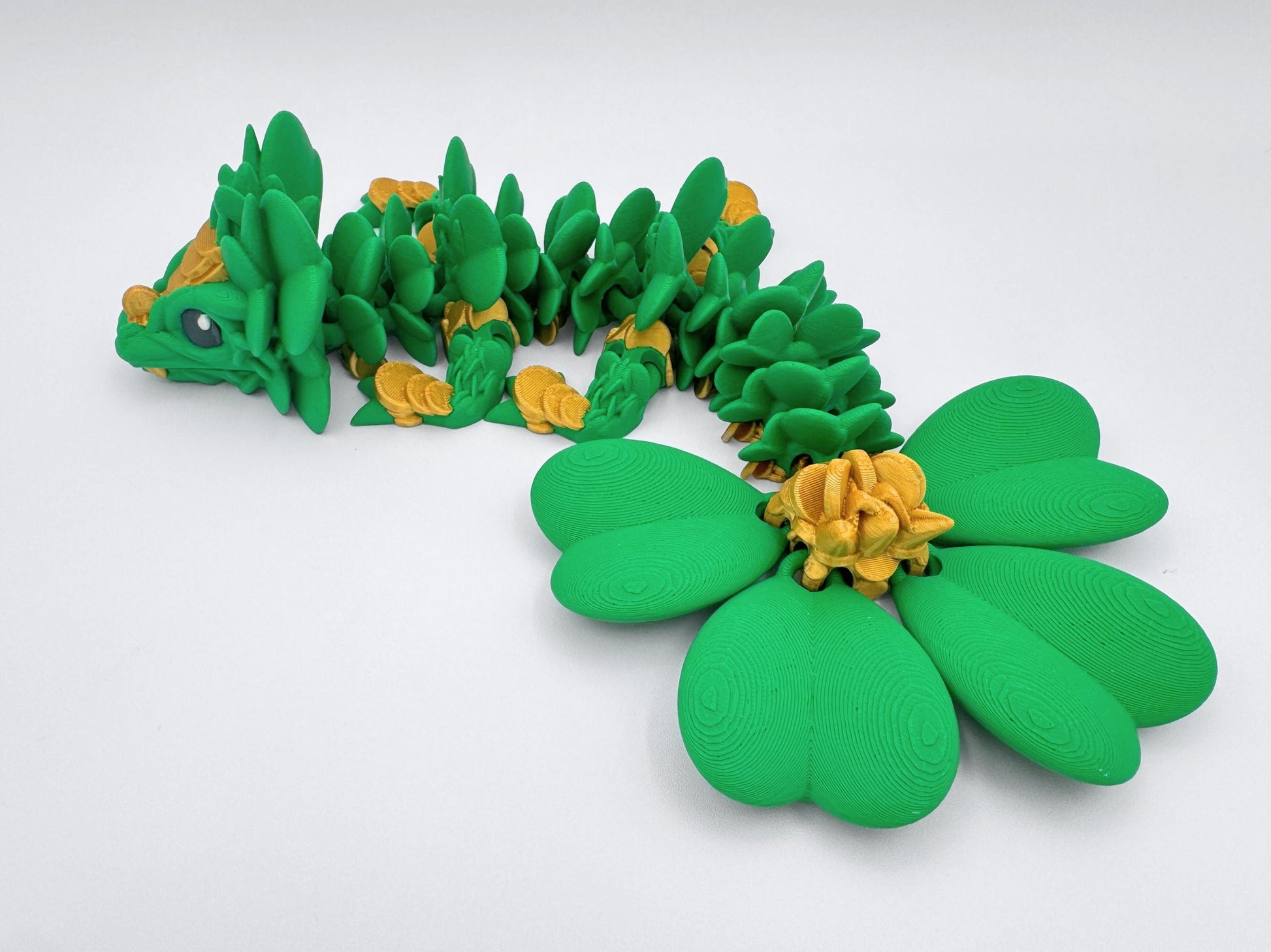 3D Printed Clover Dragon green gold and black articulated figurine with a four leaf clover tail. The dragon is made of multiple overlapping pieces that form scales. The clover is made of four large green leaves with a gold center and has a large head with a wide mouth and large eyes. Used as desk decor for seasonal gift for St. Patrick’s Day.