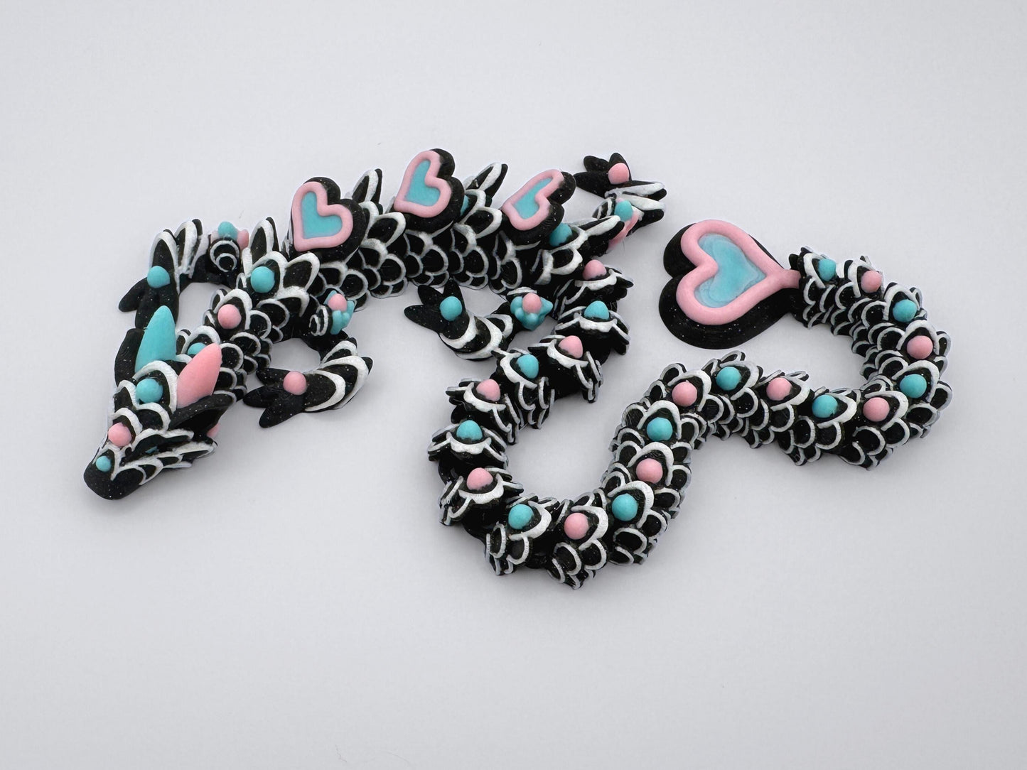 3D-printed articulated heart dragon cookie inspired fidget toy, with glitter black body, sakura pink and ice blue heart accents, and white frosting patterns. Features intricate details and a fully flexible design.