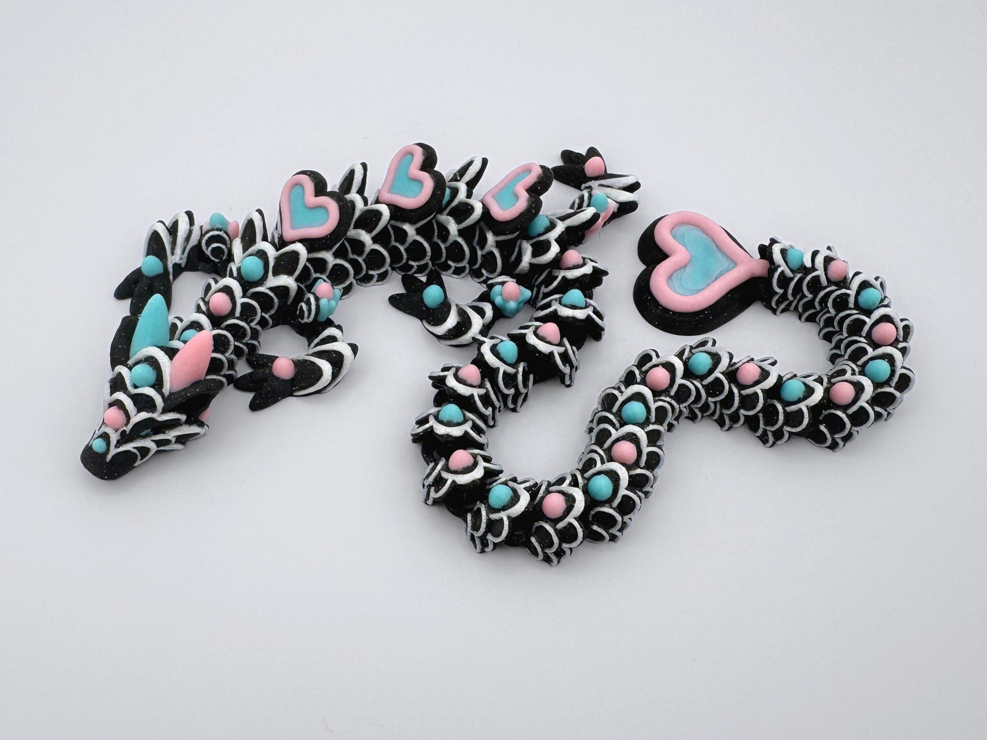 3D-printed articulated heart dragon cookie inspired fidget toy, with glitter black body, sakura pink and ice blue heart accents, and white frosting patterns. Features intricate details and a fully flexible design.
