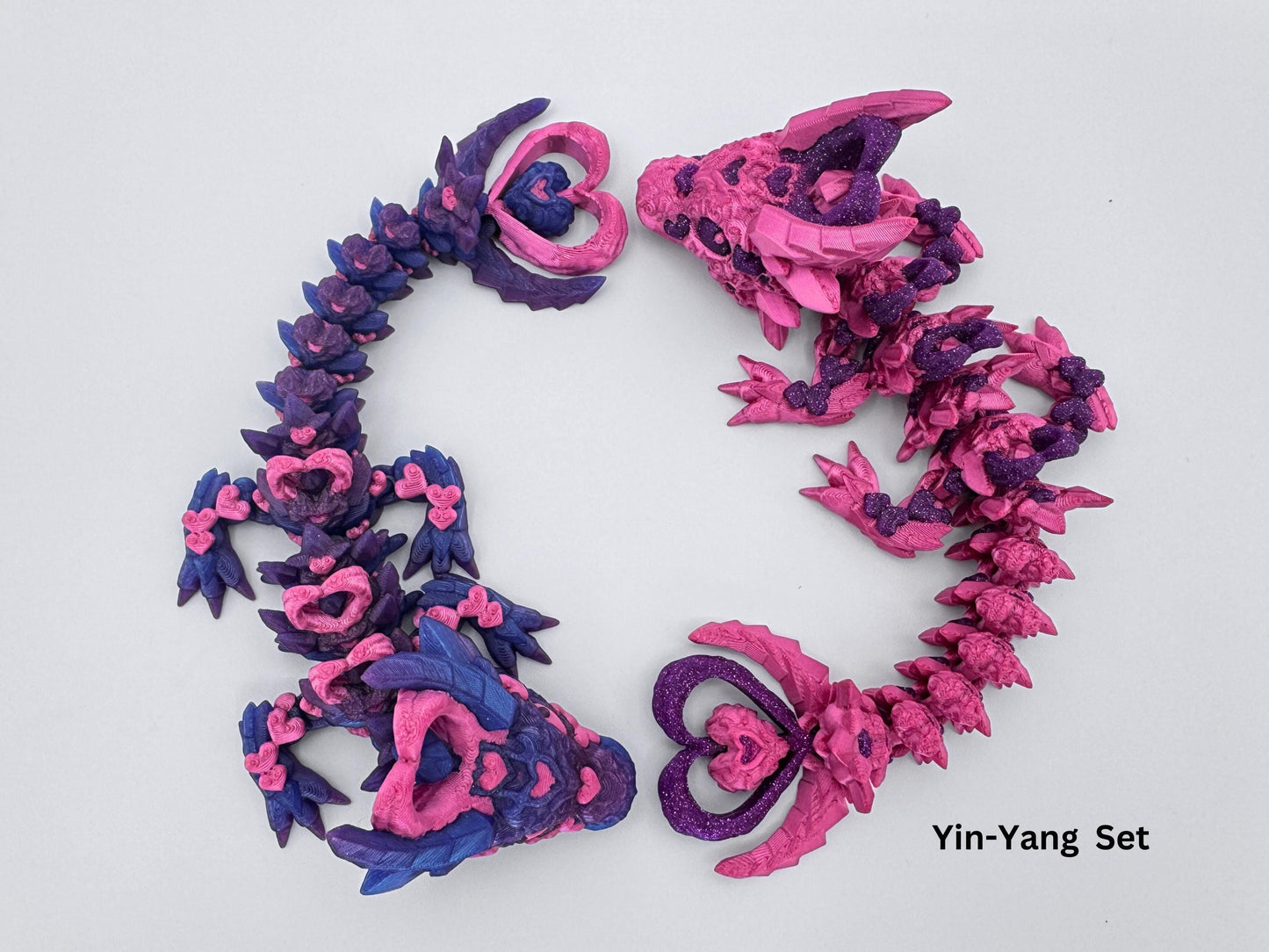 Pair of 3D-printed Dark Heart Dragon fidget toys with articulated bodies, mirroring each other in a color swap. One features a blue-to-purple gradient with magenta hearts, while the other is magenta with glitter purple hearts. Both have heart shaped horns, tail tips, and segmented flexible designs, blending fantasy and gothic aesthetics.