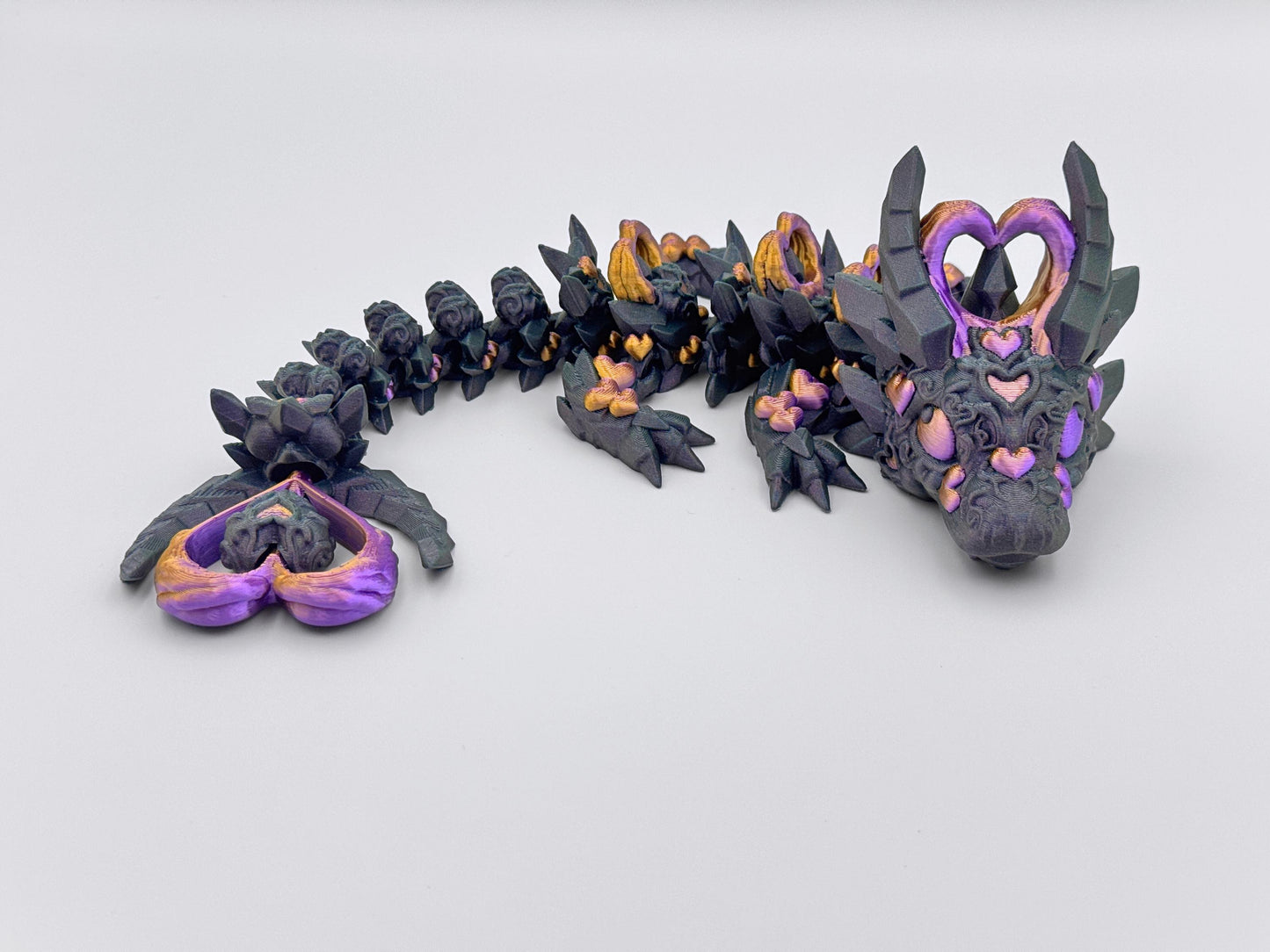3D-printed Dark Heart Dragon fidget toy with an articulated blue-black body, detailed with heart-shaped accents in gold and purple. Features heart shaped horns, a matching tail tip, and intricate sculpted textures. The segmented, flexible design allows for dynamic posing, blending gothic and fantasy aesthetics.