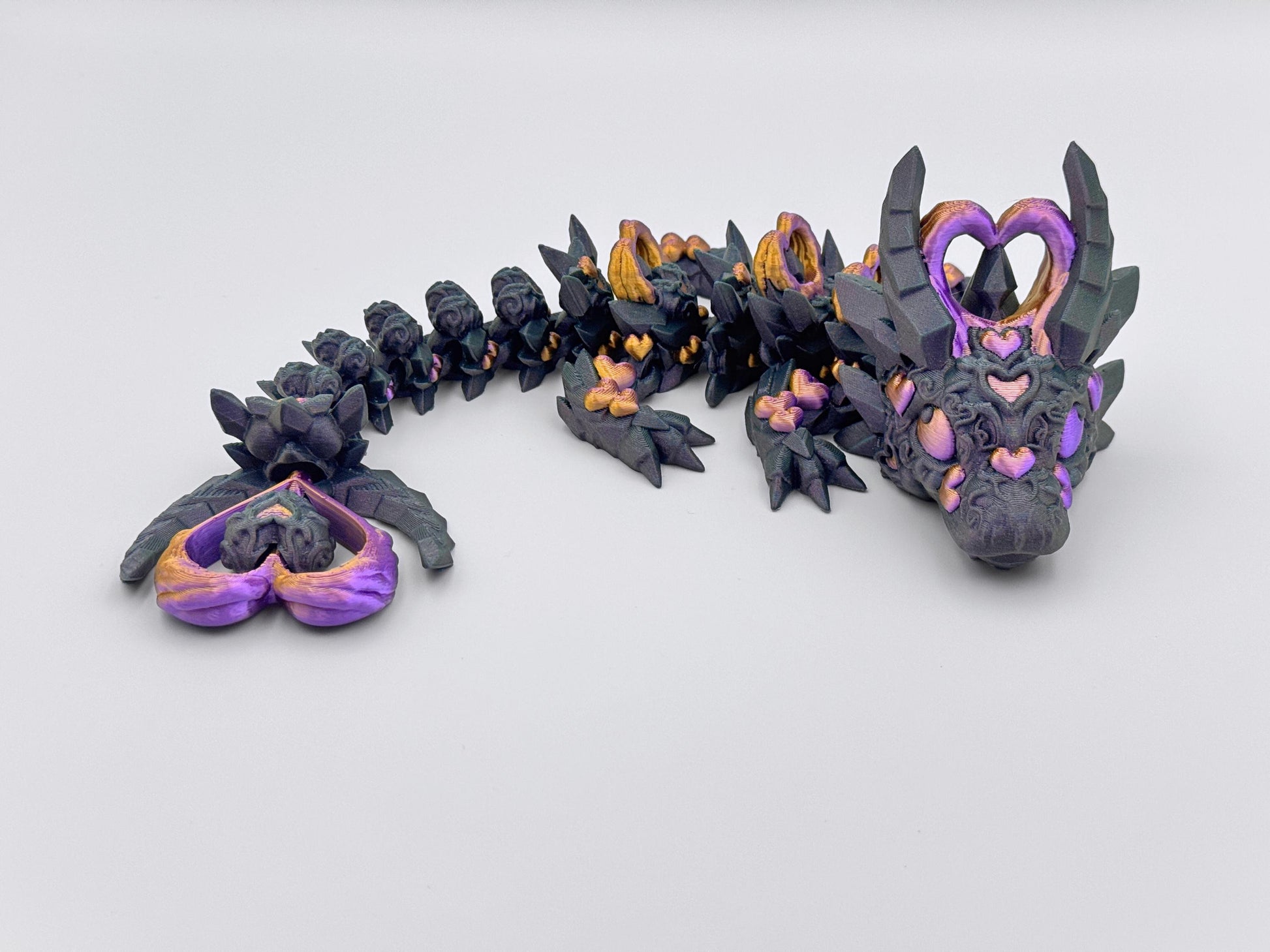 3D-printed Dark Heart Dragon fidget toy with an articulated blue-black body, detailed with heart-shaped accents in gold and purple. Features heart shaped horns, a matching tail tip, and intricate sculpted textures. The segmented, flexible design allows for dynamic posing, blending gothic and fantasy aesthetics.