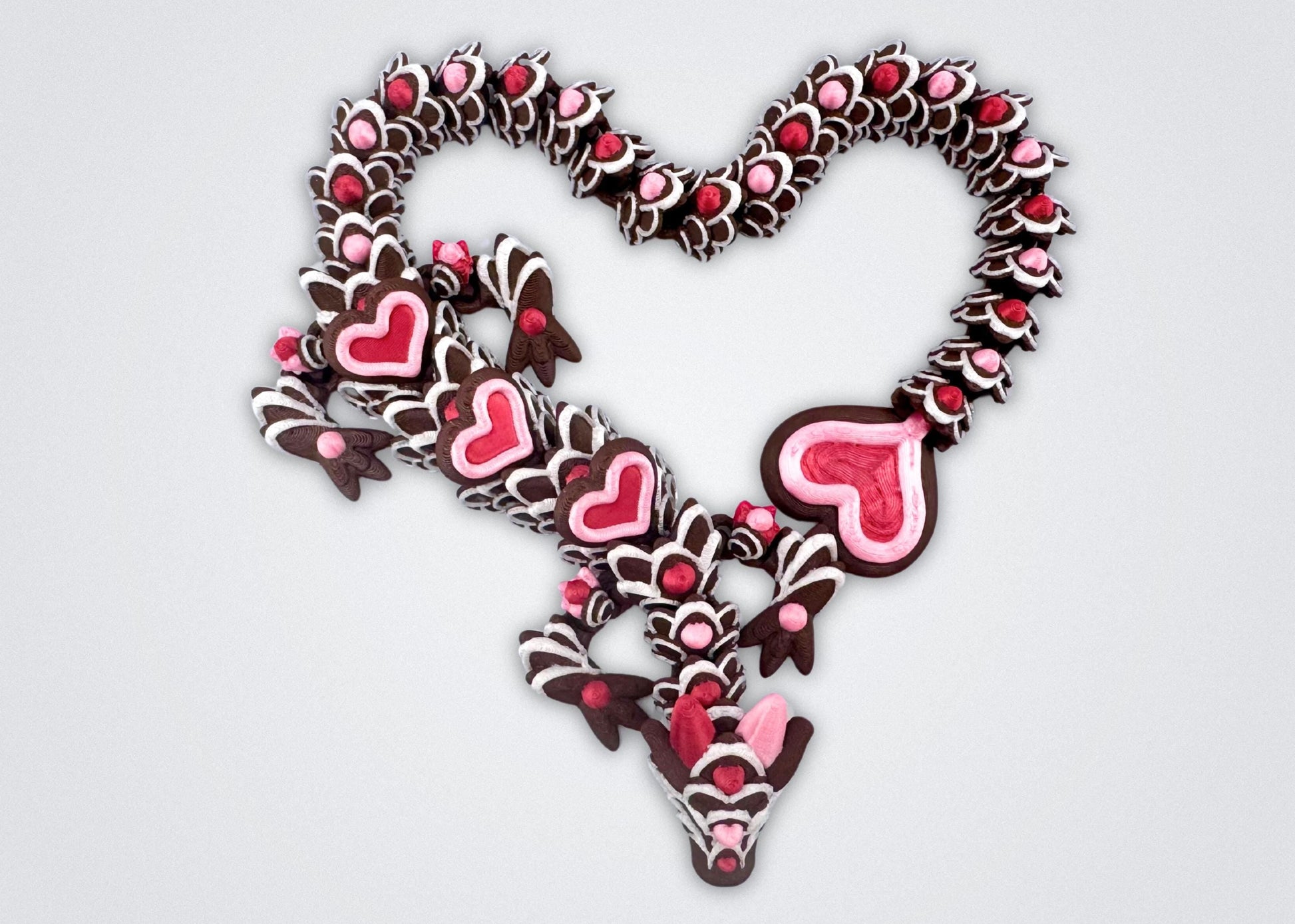 3D-printed articulated heart dragon cookie inspired fidget toy, with dark chocolate brown body, silk pink and silk red candy accents, white icing-like patterns and red hears with pink outline. Features intricate details and a fully flexible design.