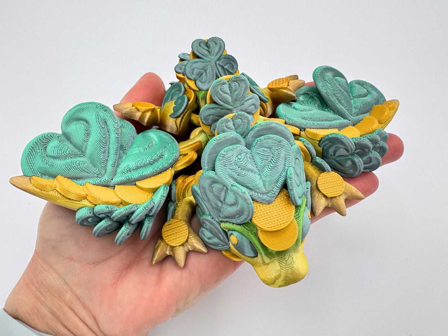 A close view of a baby clover dragon figurine cradled in a person’s hand. The dragon’s face is visible with rounded features and layered clover leaves along the head, wings and back. Its body appears articulated and folded slightly inward, giving a clear view of the scale and detail of the figure in the palm.