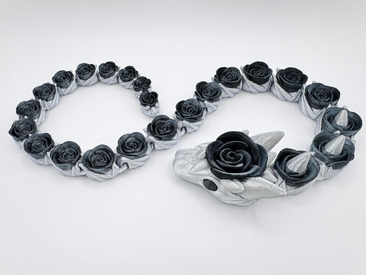 Black and silver 3D printed rose snake with fully articulated segments arranged in a smooth curve. Each body segment is shaped like a detailed black rose set into a silver coil. The head features pointed silver horns and a central rose bloom. Displayed on a white background to show the full body and gothic floral details of the figurine.