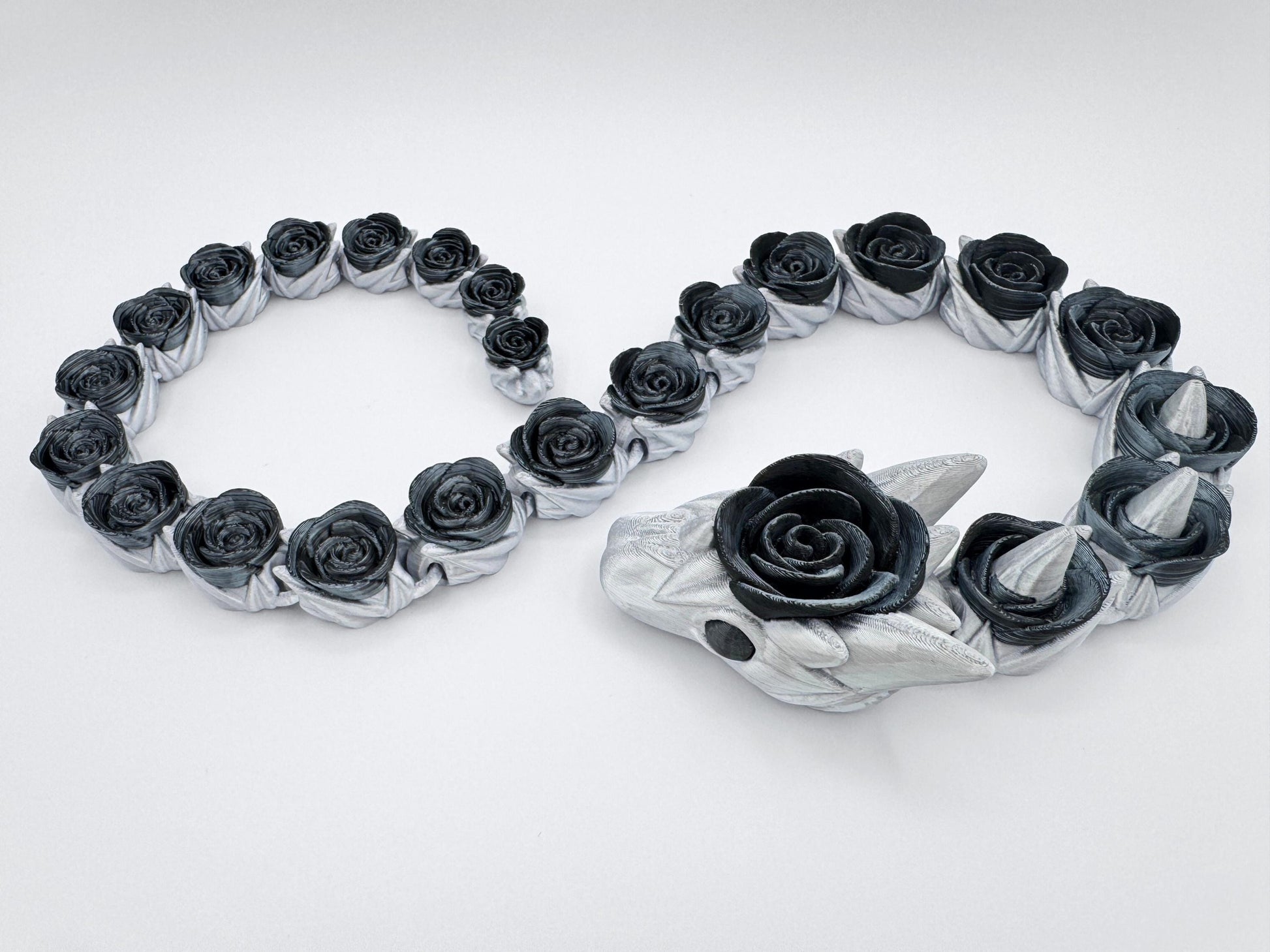 Black and silver 3D printed rose snake with fully articulated segments arranged in a smooth curve. Each body segment is shaped like a detailed black rose set into a silver coil. The head features pointed silver horns and a central rose bloom. Displayed on a white background to show the full body and gothic floral details of the figurine.