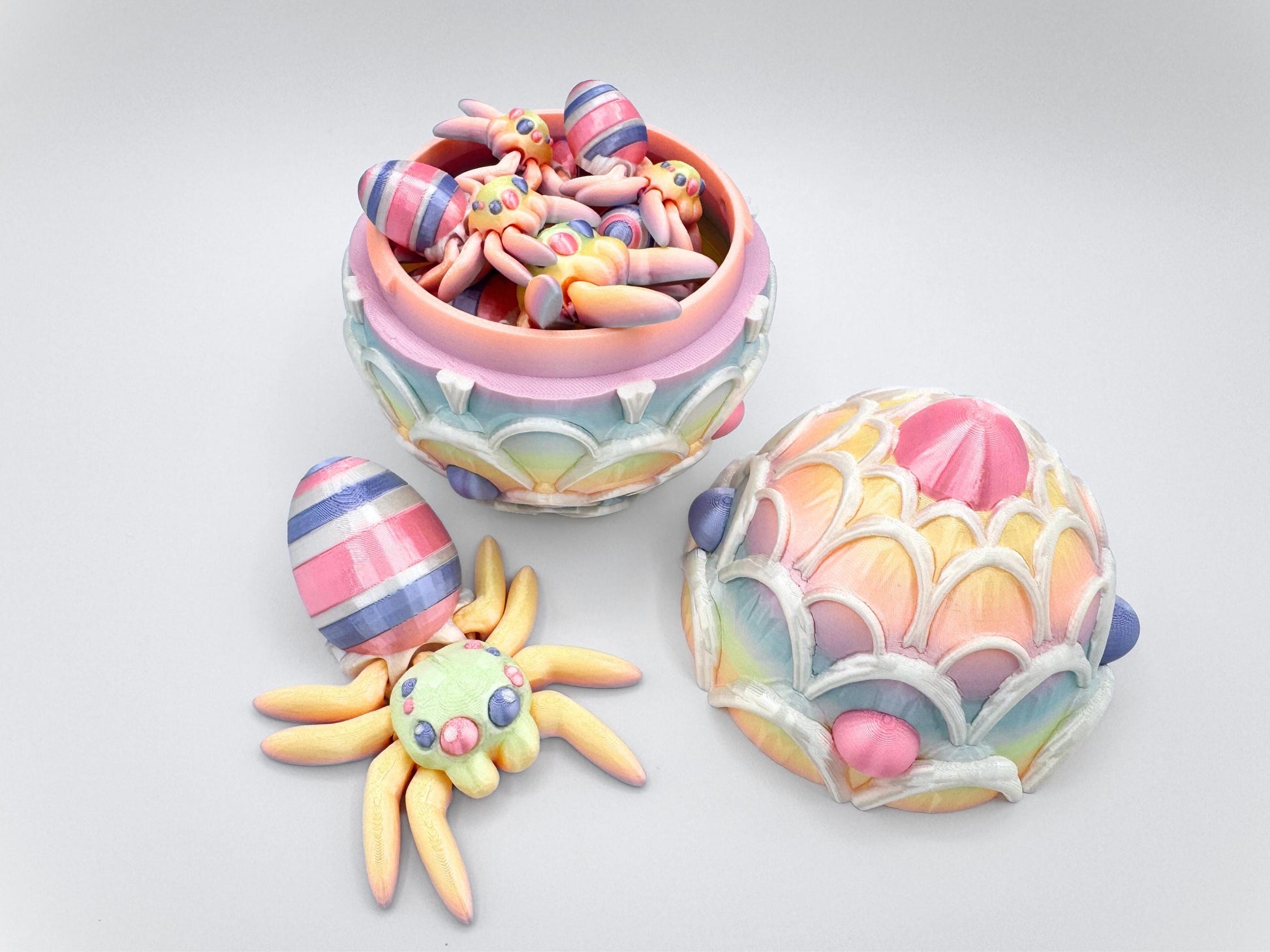 3D-printed Easter Egg Spider with a soft rainbow gradient body, seamlessly blending pastel tones. The egg on its back features bold pink, violet, and white stripes, adding a festive touch. The large matching egg is decorated with white icing and pink and violet candy accents, serving as both a decorative piece and a storage container for Easter stuffers.
