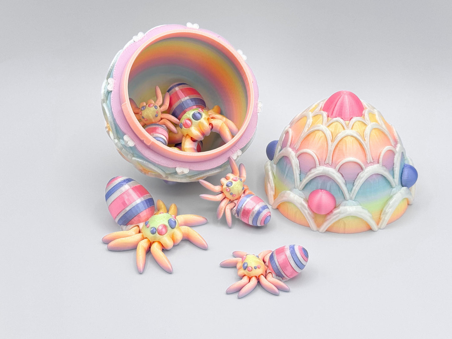 3D-printed Easter Egg Spider with a soft rainbow gradient body, seamlessly blending pastel tones. The egg on its back features bold pink, violet, and white stripes, adding a festive touch. The large matching egg is decorated with white icing and pink and violet candy accents, serving as both a decorative piece and a storage container for Easter stuffers.