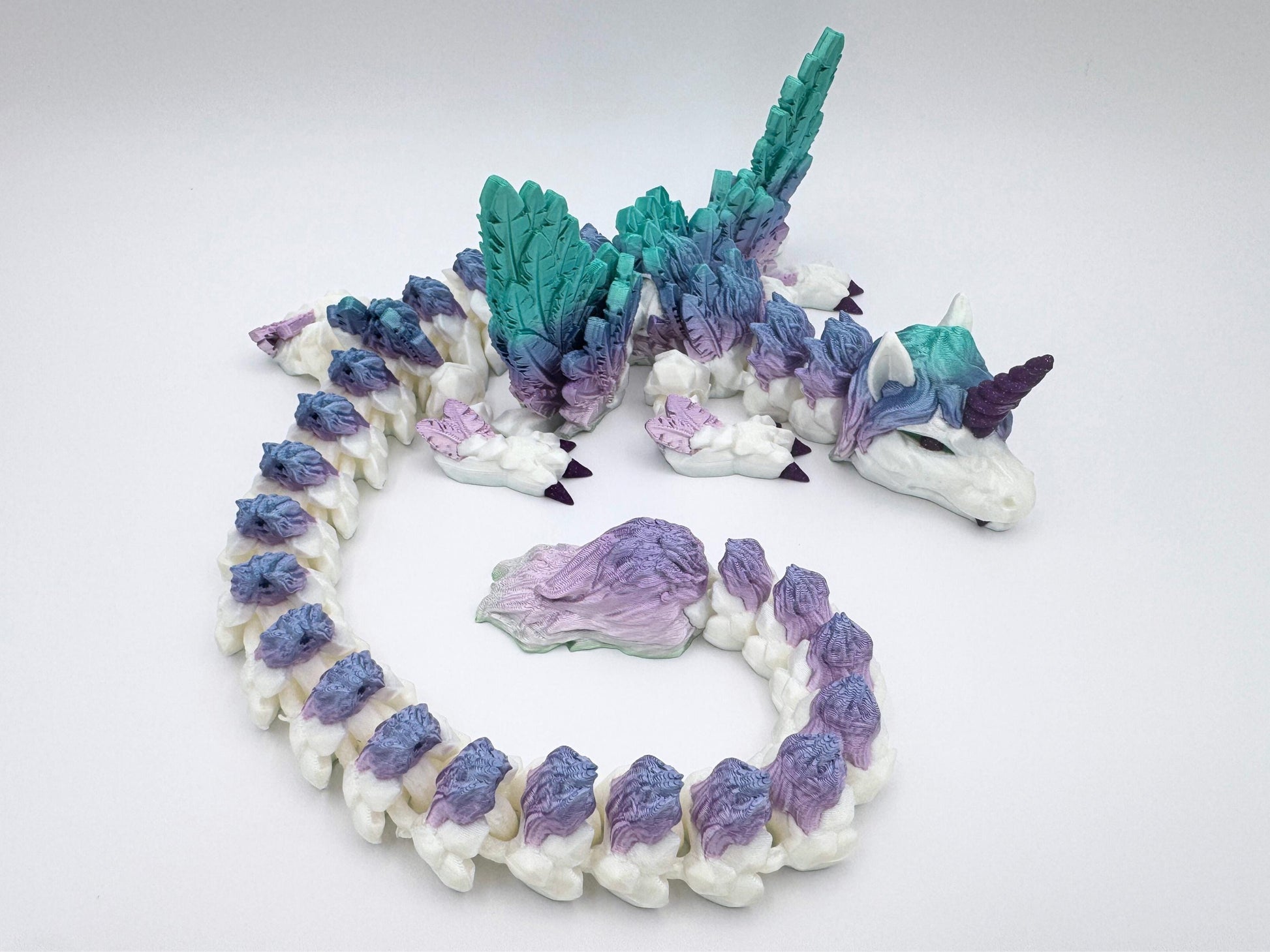 3D printed unicorn dragon lying on a flat white surface. The dragon has a unicorn horn and is curled up in a sleeping position. Features a fully articulated segmented body with silk rainbow filament in teal, mint and lavender. Large feathered wings extend from its back. The dragon has a sculpted mane, deep purple unicorn horn and matching purple claws. Tail ends in a detailed plume of feathery texture. Printed with glossy finish and visible scale detailing.