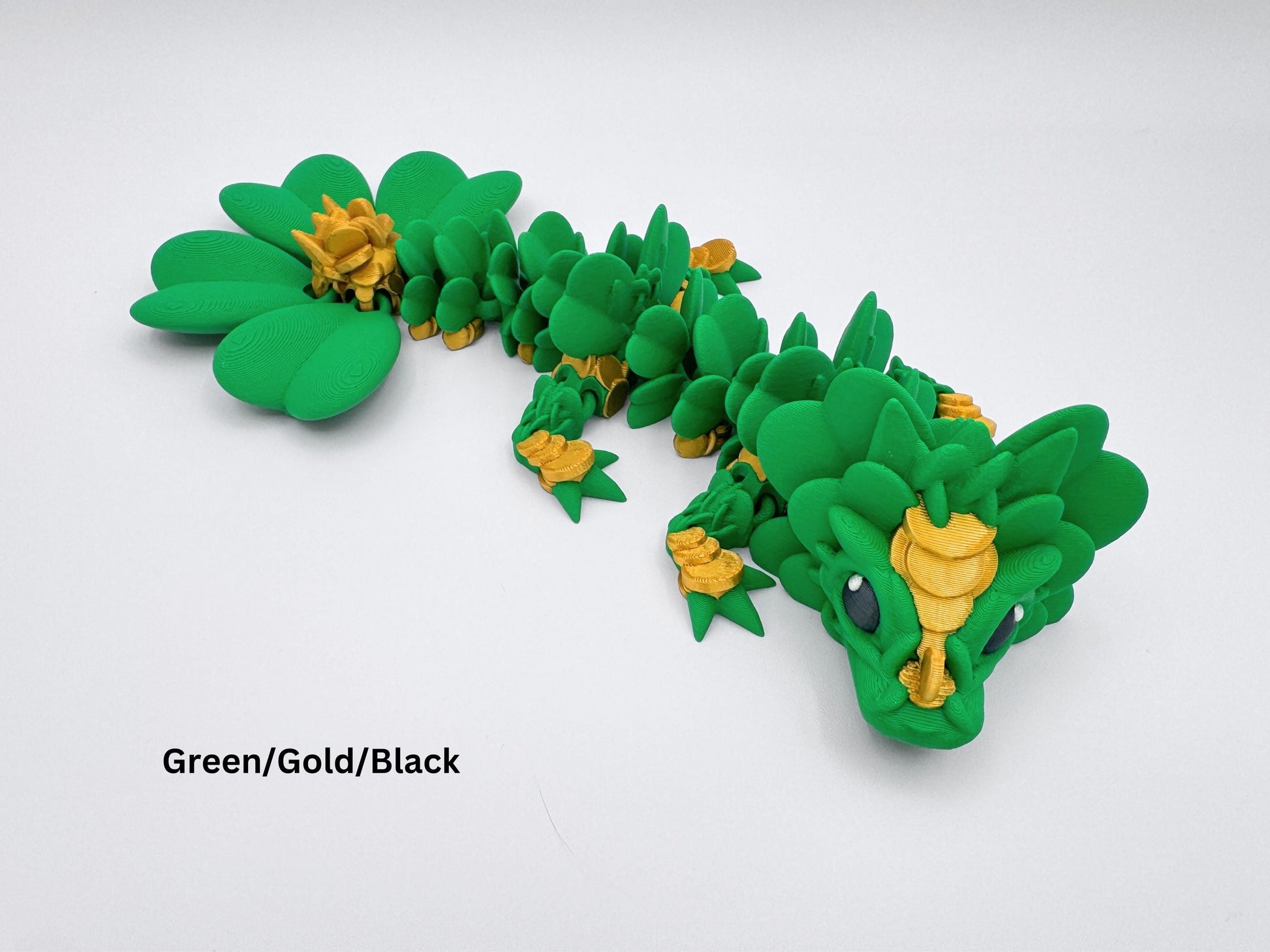 3D Printed Clover Dragon green gold and black articulated figurine with a clover 4 leaf design along its body. The dragon is made up of many small, overlapping leaves and has a large head with a wide mouth and large eyes. The dragon's tail is curled up behind it and is lying on a white surface.  Used as desk decor for seasonal gift for St. Patrick’s Day.