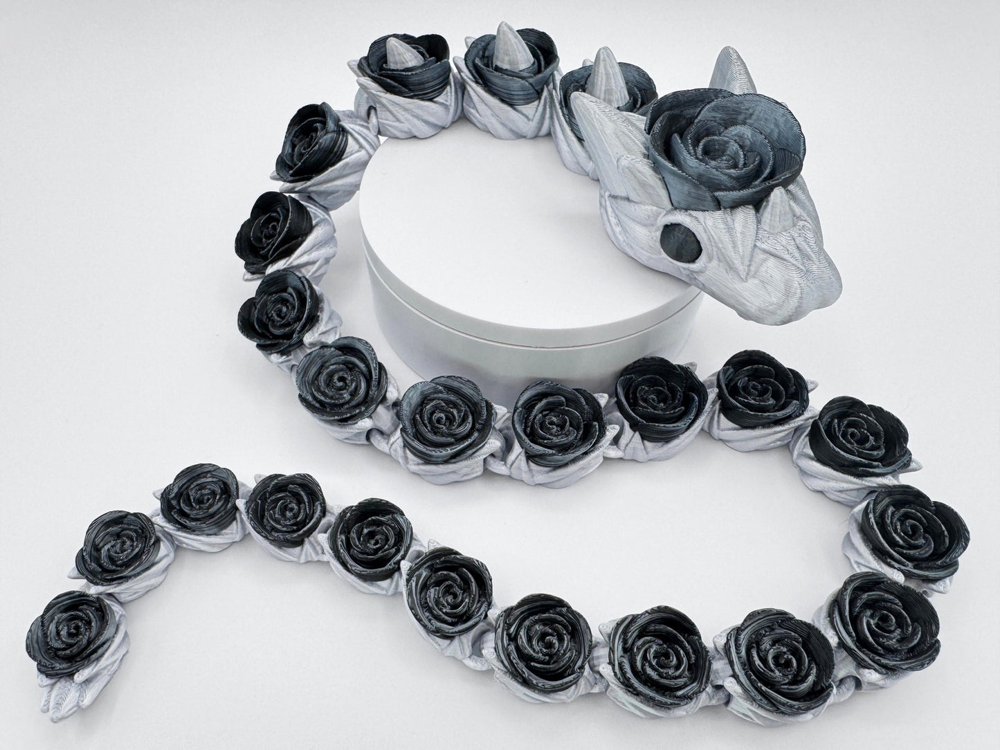 Black roses snake draped around a white circular pedestal, showing off its full articulated length. The dark rose blooms wrap around the silver body, with detailed scales, horns, and flower textures visible. Posed in motion to highlight its flexible display potential.