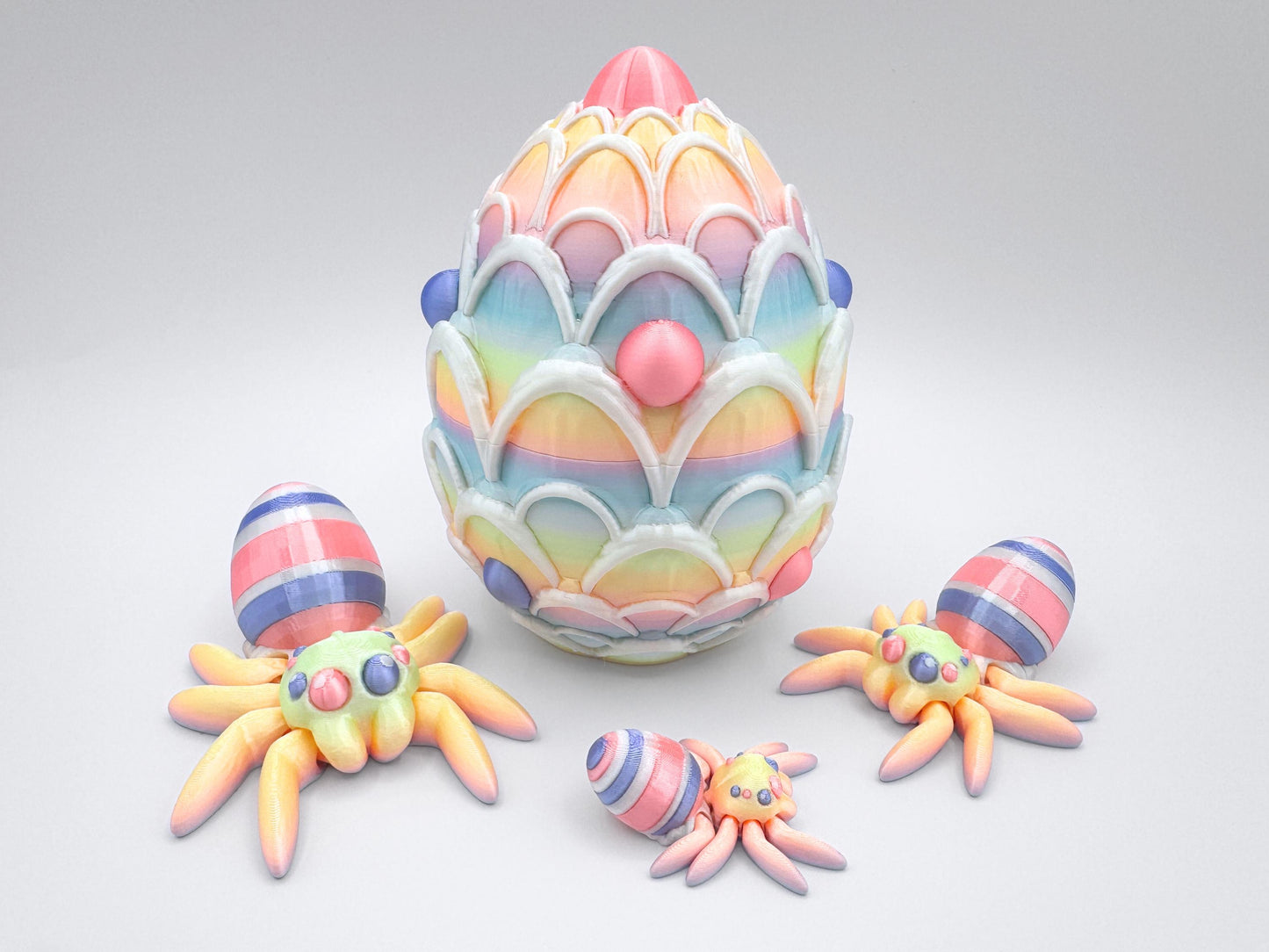 3D-printed Easter Egg Spider with a soft rainbow gradient body, seamlessly blending pastel tones. The egg on its back features bold pink, violet, and white stripes, adding a festive touch. The large matching egg is decorated with white icing and pink and violet candy accents, serving as both a decorative piece and a storage container for Easter stuffers.