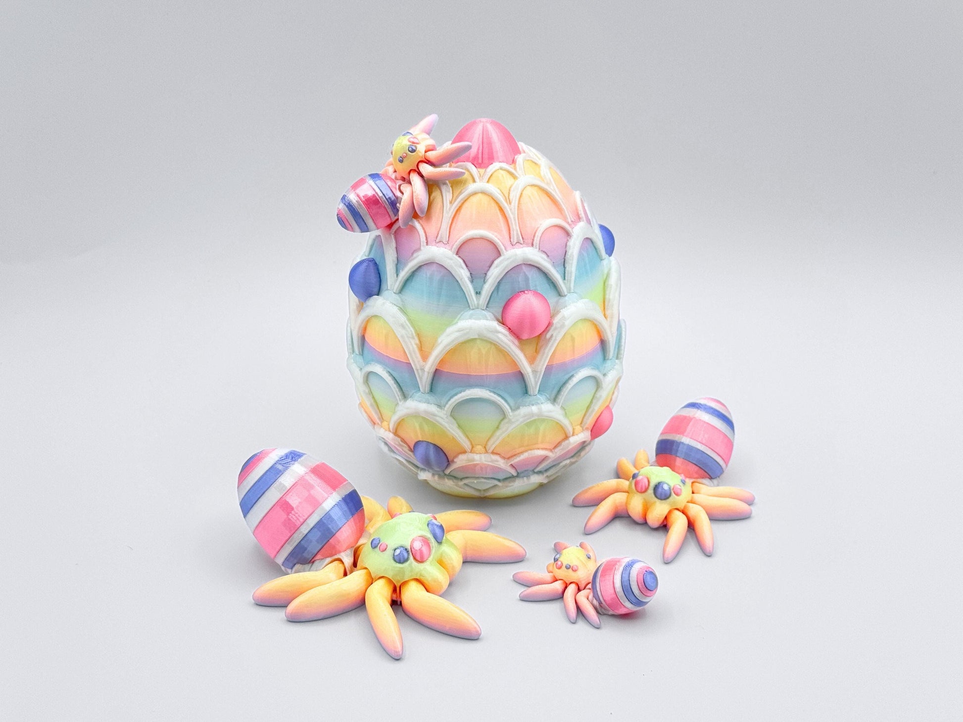 3D-printed Easter Egg Spider with a soft rainbow gradient body, seamlessly blending pastel tones. The egg on its back features bold pink, violet, and white stripes, adding a festive touch. The large matching egg is decorated with white icing and pink and violet candy accents, serving as both a decorative piece and a storage container for Easter stuffers.