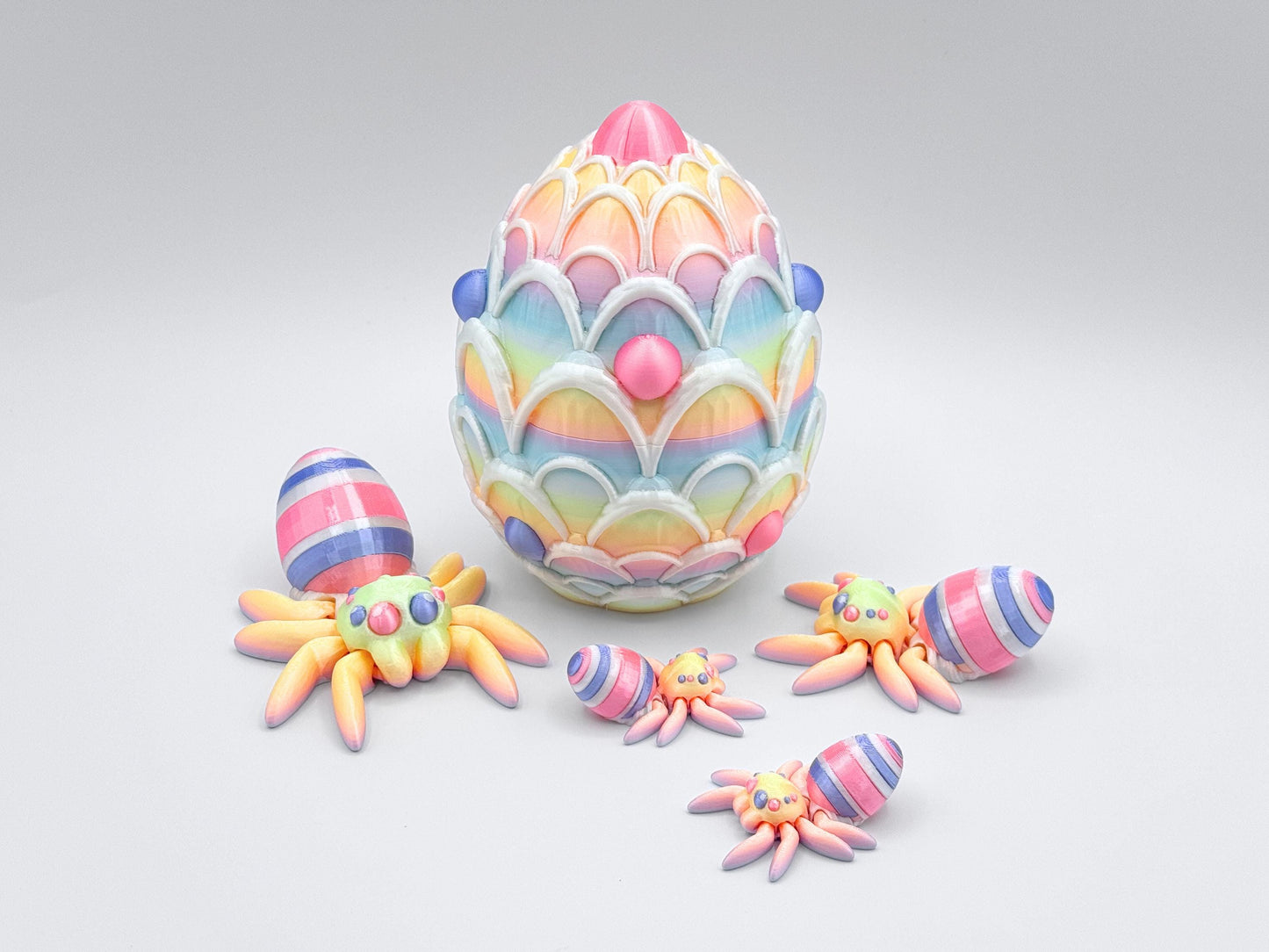 3D-printed Easter Egg Spider with a soft rainbow gradient body, seamlessly blending pastel tones. The egg on its back features bold pink, violet, and white stripes, adding a festive touch. The large matching egg is decorated with white icing and pink and violet candy accents, serving as both a decorative piece and a storage container for Easter stuffers.