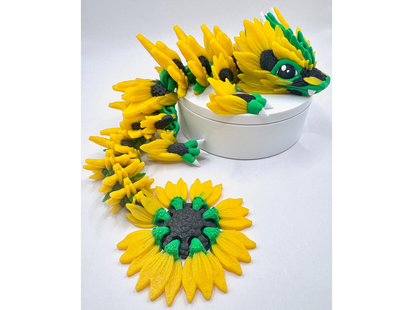 A green and yellow 3D printed dragon figurine with sunflower inspired petals. The dragon is laying on a white round platform with its tail curled around the platform. The tail has sunflower shaped details. The dragon's head is facing the right side of the image and has green eyes and a black nose.