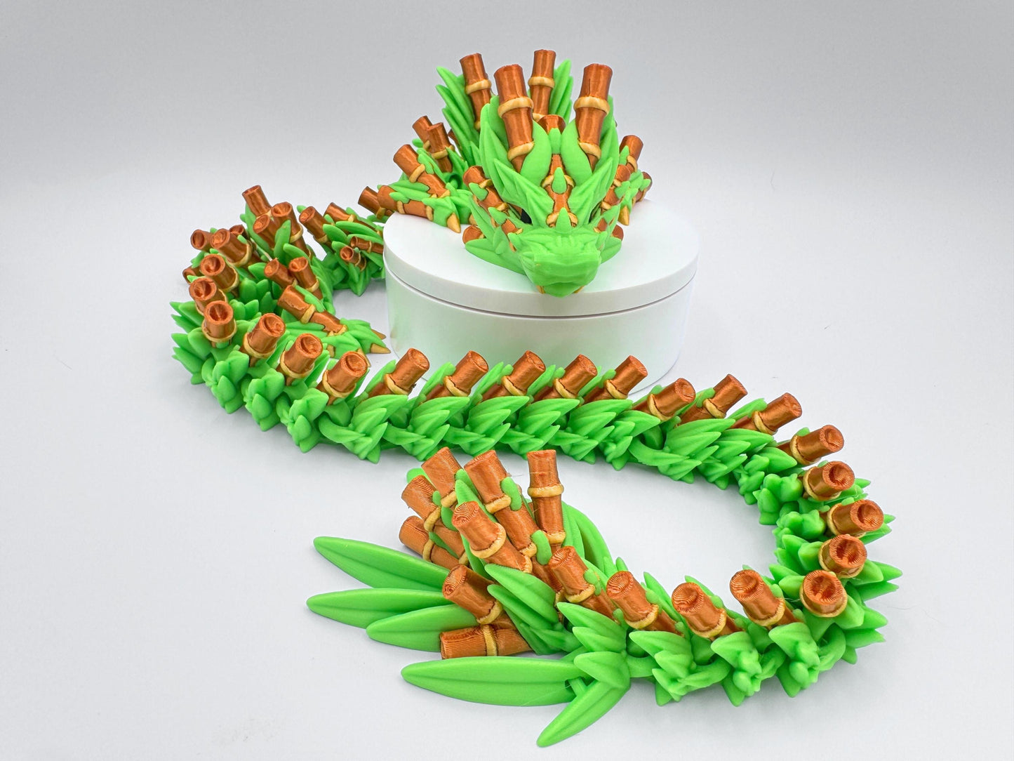 3D printed articulated Bamboo Dragon posed around a white round stand. The long, flexible body features bright green leaf segments and bamboo-gold stalk details, with the head framed by layered leaves. The figure curves in an S shape to show poseable links and texture. Neutral white background keeps focus on the dragon’s color contrast and articulation.