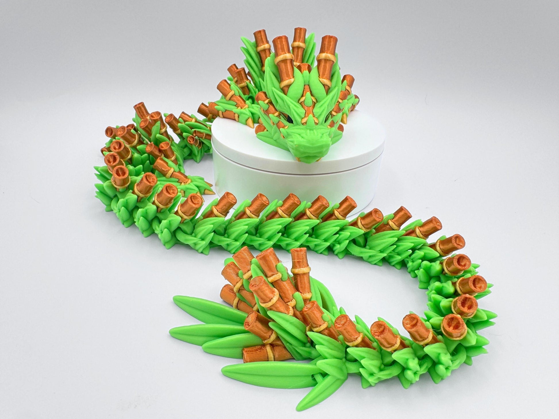 3D printed articulated Bamboo Dragon posed around a white round stand. The long, flexible body features bright green leaf segments and bamboo-gold stalk details, with the head framed by layered leaves. The figure curves in an S shape to show poseable links and texture. Neutral white background keeps focus on the dragon’s color contrast and articulation.