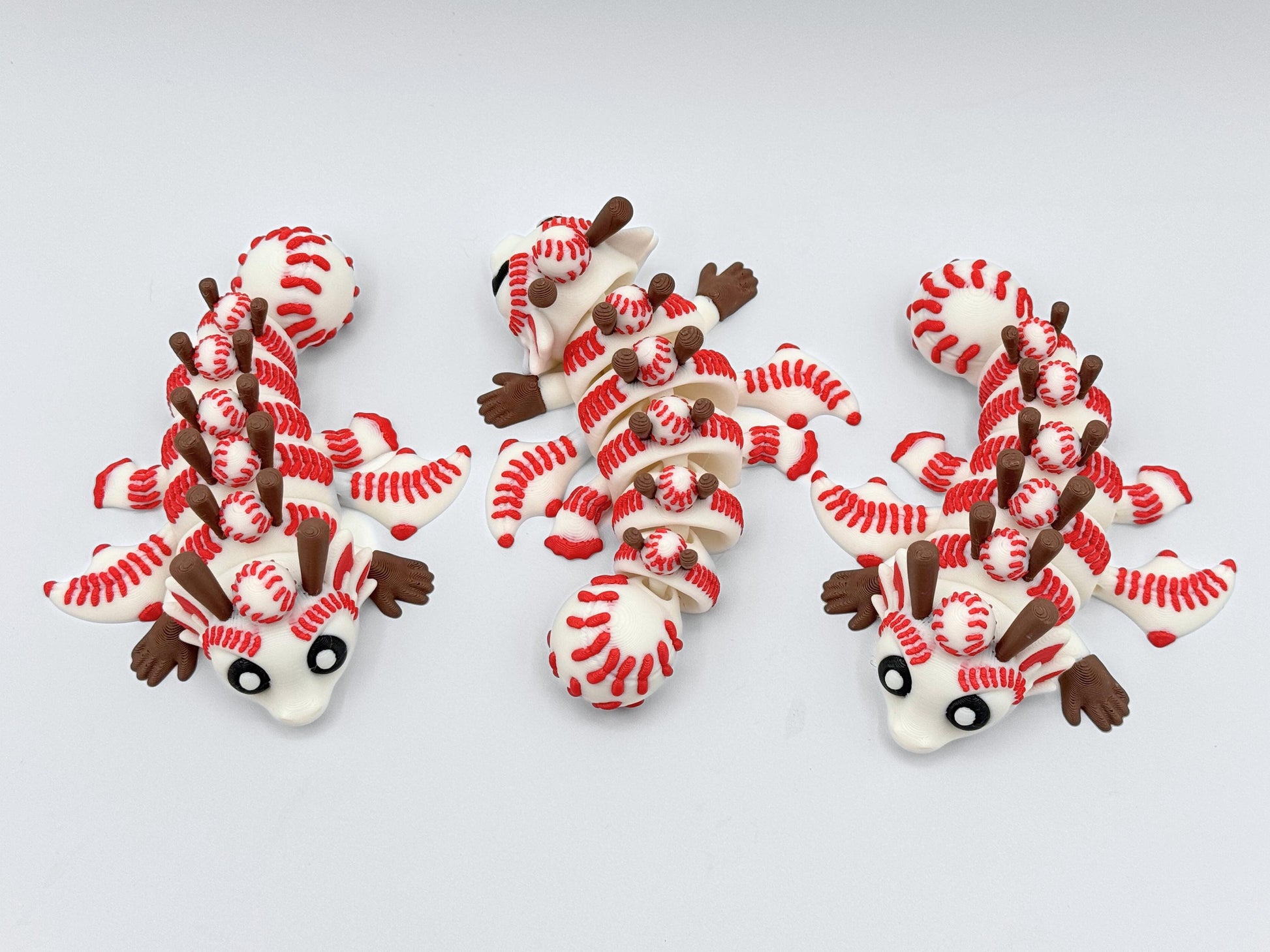 Three articulated Baseball Dragon figurines laid out in different positions, showcasing flexible segmented bodies, white and red baseball themed textures, brown limbs, and oversized eyes.