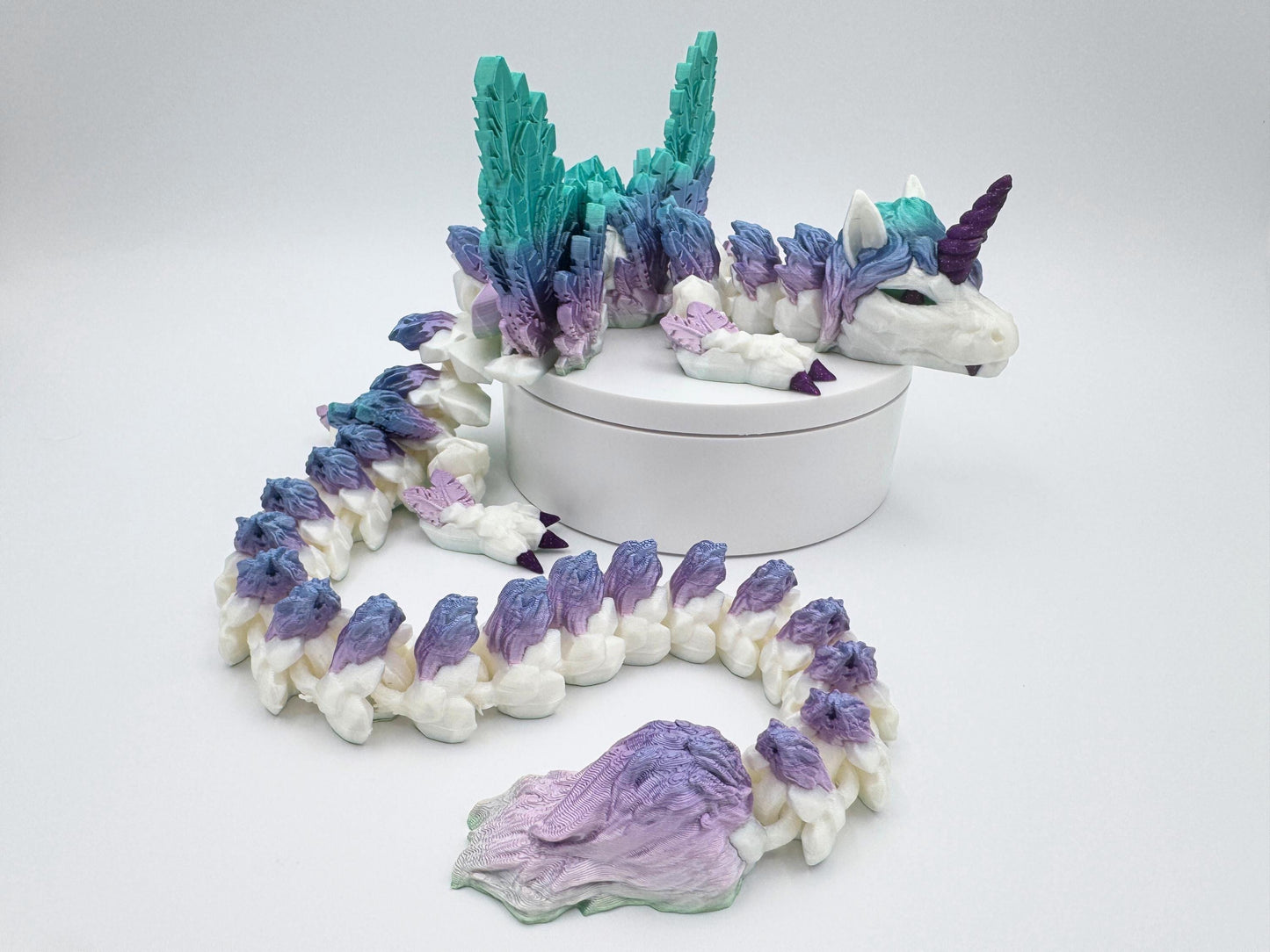 3D printed unicorn dragon curled on a round white platform. Printed in silk rainbow filament with bright teal, aqua, lavender and white colors. Large feather textured wings rise from the back. The dragon has a deep purple spiral horn, purple claws and a flowing mane and tail in blended bright rainbow tones. Its articulated body coils naturally around the base. Sculpted details include feathers, scales, and a smooth, glossy shimmer finish.
