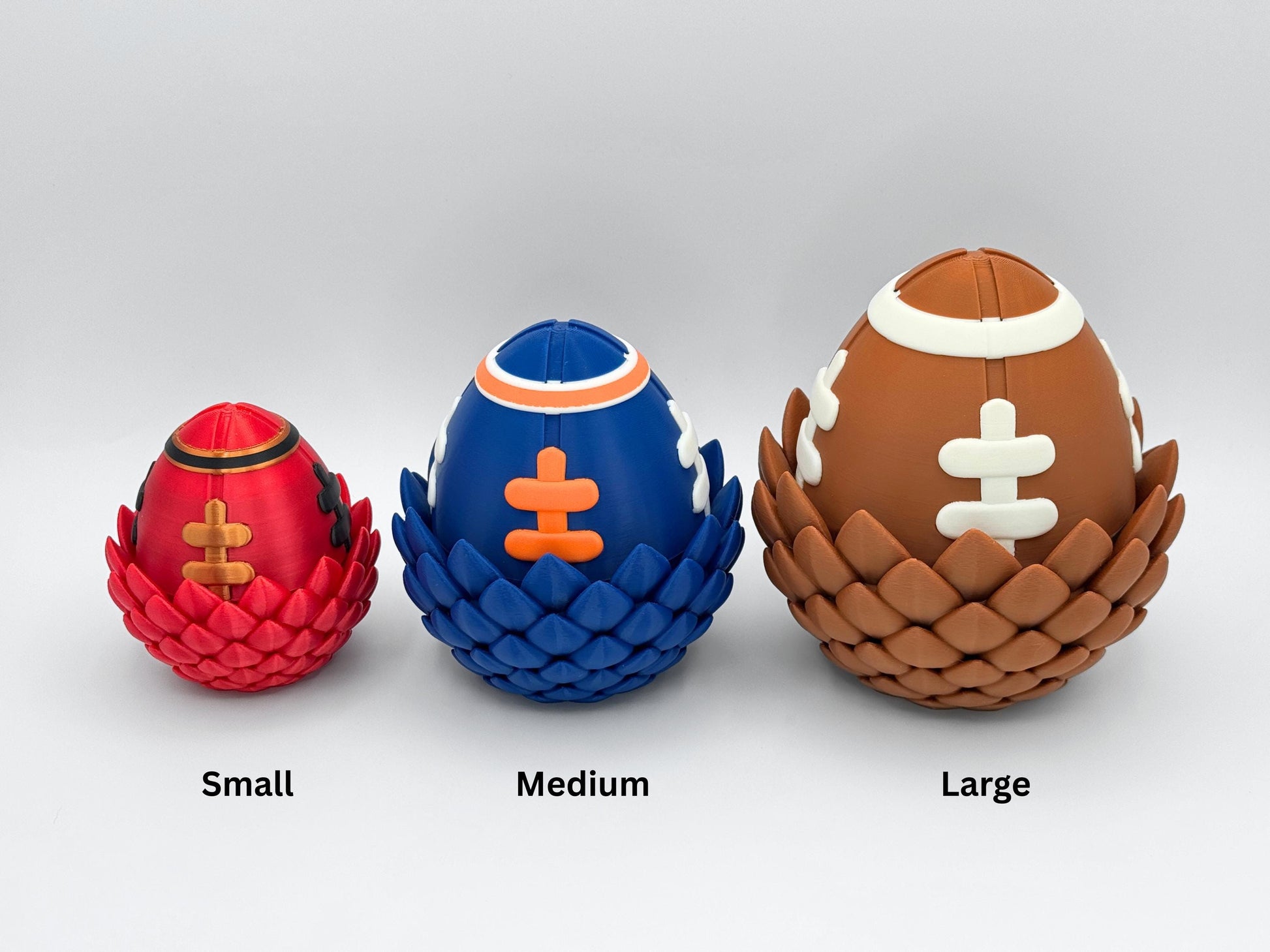 Small red and gold, medium blue and orange, and large brown and white football dragon eggs side by side for size comparison. Each 3D printed egg has a textured scale base and football lace pattern, representing different team-inspired color themes.
