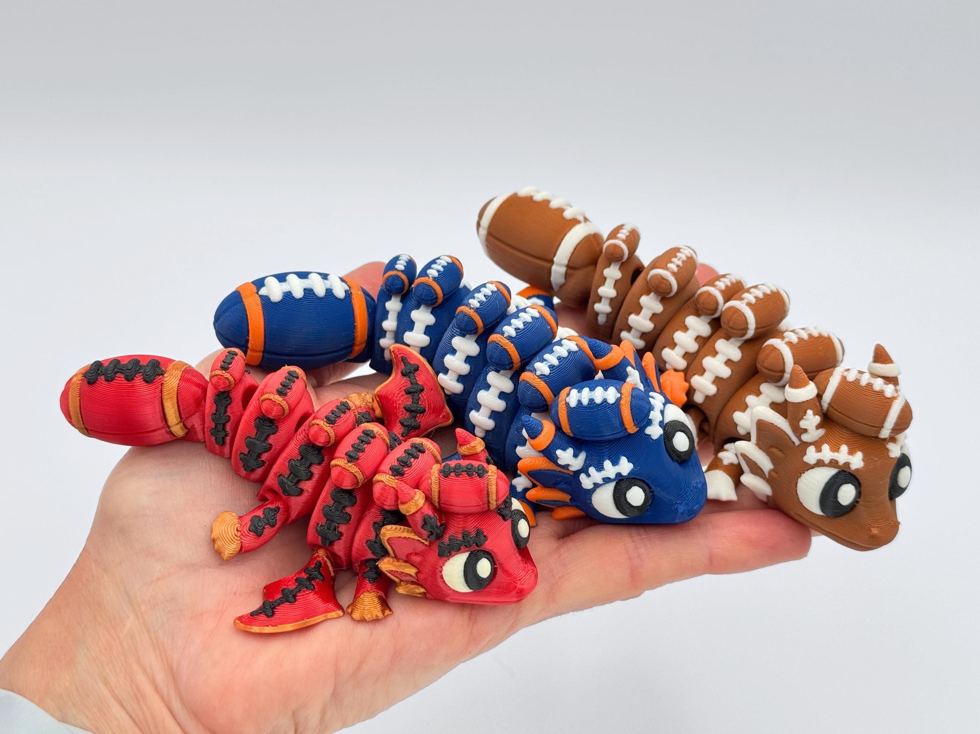 Hand holding three articulated football dragons in red and gold, blue and orange, and brown and white, showing smooth 3D printed finish, football lace detailing and flexible segmented design.